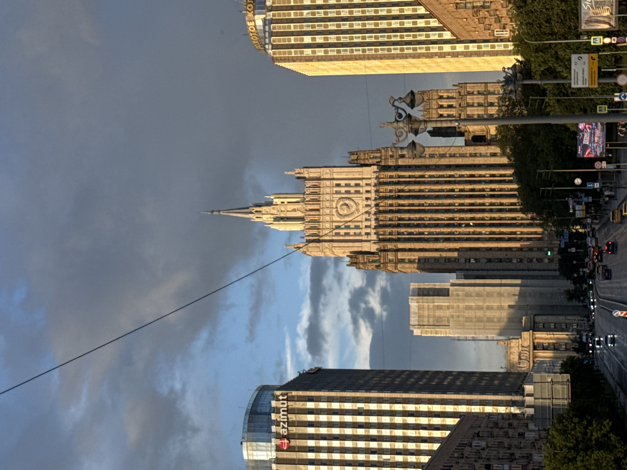 The image shows a street view in Moscow, Russia, featuring several prominent buildings and urban elements under a cloudy sky. The central and most striking structure is the Stalinist skyscraper, the Ministry of Foreign Affairs building, with its distinctive spire and ornate facade. To the left, a modern glass-fronted building with the "azimut" logo is visible. To the right, another tall building, likely the "Zolotoye Koltso" hotel, stands with its facade illuminated by the sun. A street with cars driving on it occupies the lower part of the image, lined with trees and streetlights. Several billboards and traffic signs are also present along the road. The sky is dramatic, with dark clouds interspersed with patches of blue and sunlight breaking through. The overall scene depicts a busy urban environment with a mix of architectural styles, from historical to contemporary.