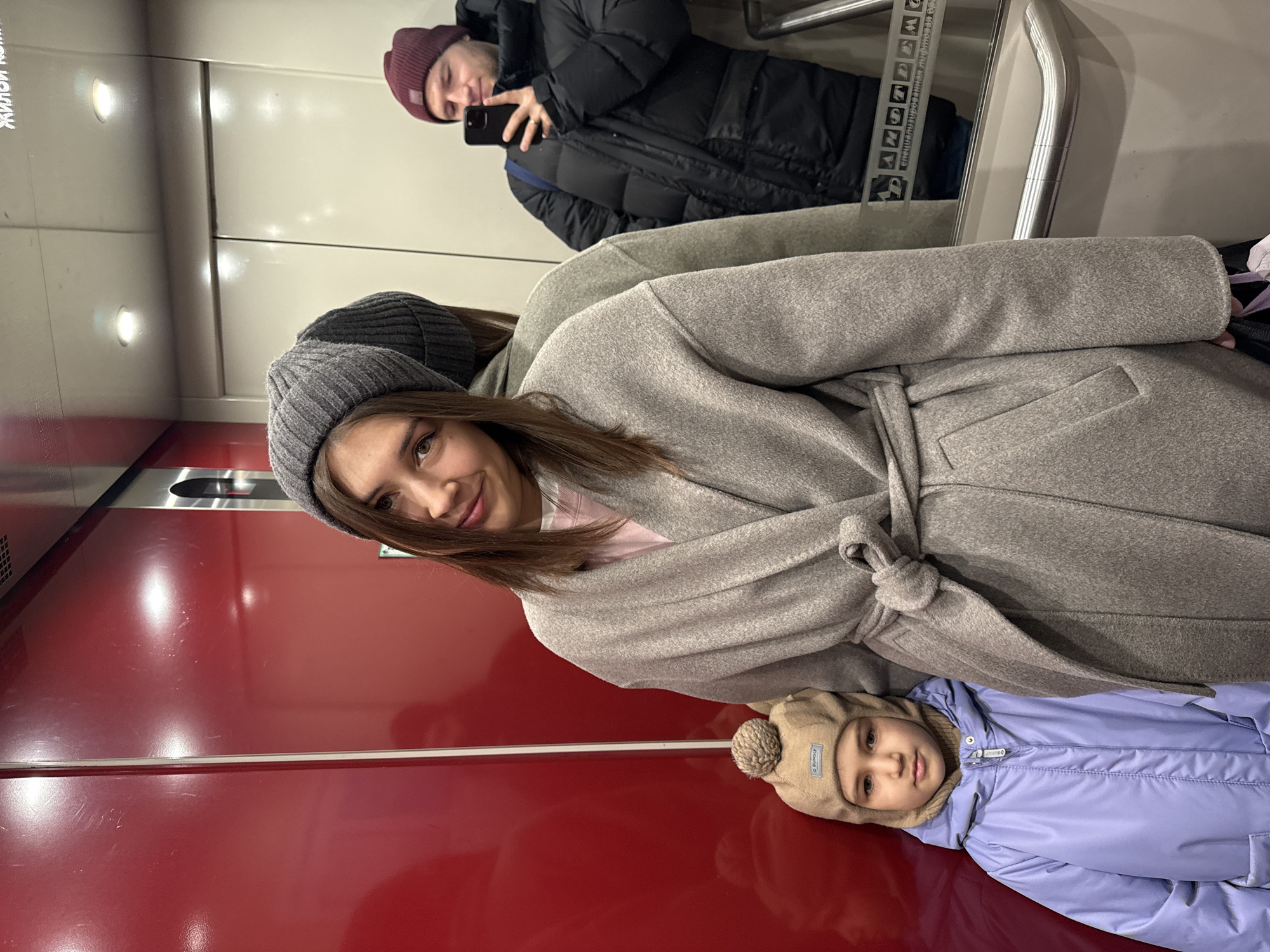 A medium shot shows three people in an elevator in Krasnoyarsk, Russia. In the bottom center of the image, a young child wearing a tan hat and blue jacket lies on the red elevator floor. A woman wearing a grey hat and matching grey coat is centrally located above the child, looking at the camera. In the background, the reflection in the elevator mirror shows a man wearing a dark-colored jacket and burgundy knit cap taking a photo with a phone. The elevator has white upper walls and a red lower section.
