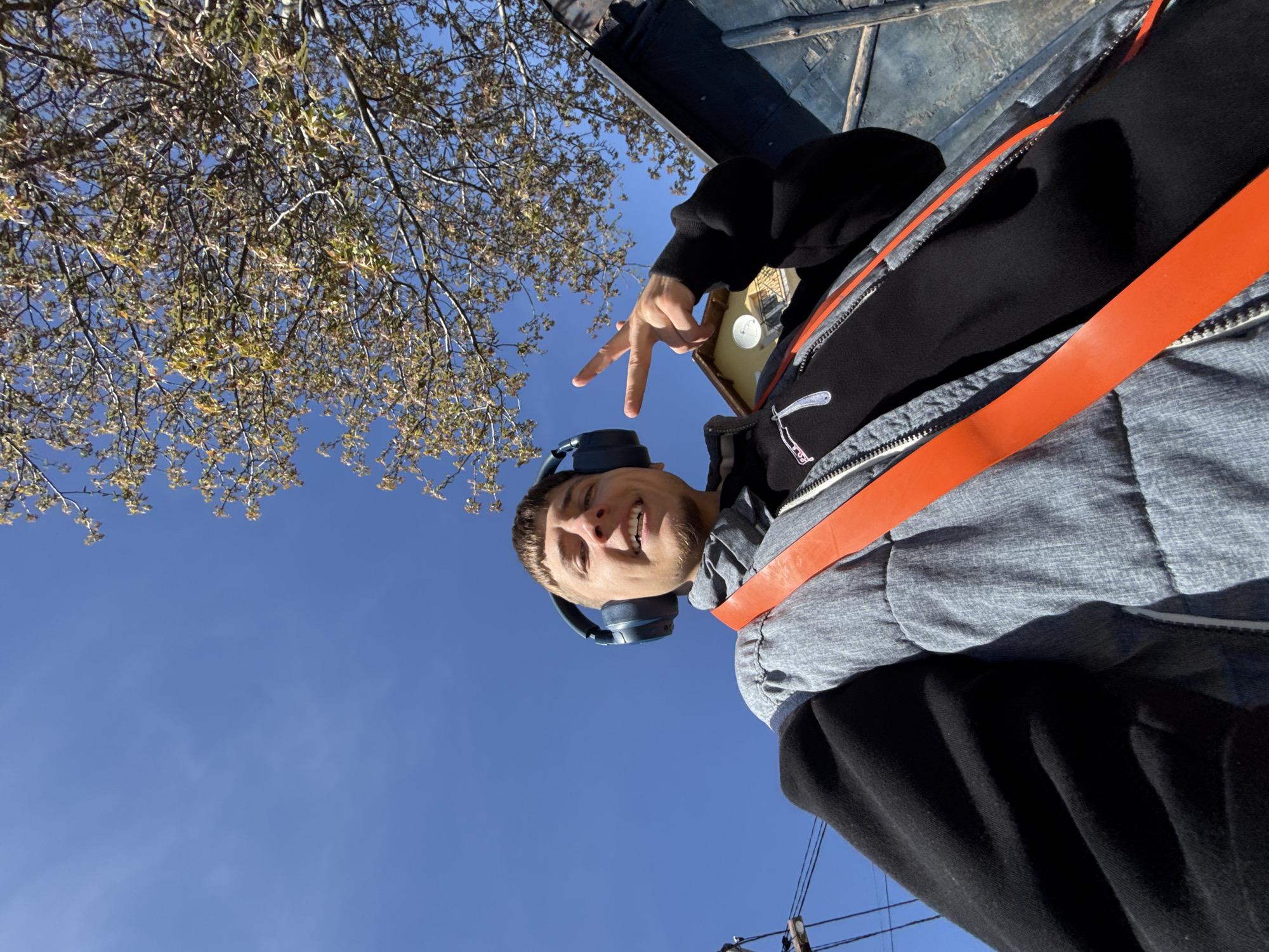 A young man is captured in a low-angle, rotated selfie-style photograph, taken outdoors in what appears to be Buhovo, Bulgaria. The image is oriented sideways, with the sky dominating the background.
The primary subject is a smiling young man, looking directly at the camera. He has short hair and a light beard, and is wearing blue over-ear headphones. He wears a grey hooded jacket over a black long-sleeved shirt, which features an embroidered design resembling a straight razor on the left chest area. A prominent orange strap, possibly from a backpack or a safety harness, crosses diagonally over his chest. He is holding his left hand up, making a "peace" sign (V-sign) with his index and middle fingers. His expression is one of clear happiness or joy, indicated by his wide smile showing teeth.
The setting is outdoors on a bright, sunny day, suggested by the clear, vibrant blue sky that fills most of the background. In what would be the upper-left of a correctly oriented image, tree branches with light green or yellow budding leaves are visible, hinting at spring or early summer. A dark, possibly wooden or metallic structure is partially visible in the upper right. Thin power lines or cables are also faintly seen against the sky in the lower portion of the frame. No discernible text is visible in the image, other than the razor symbol.