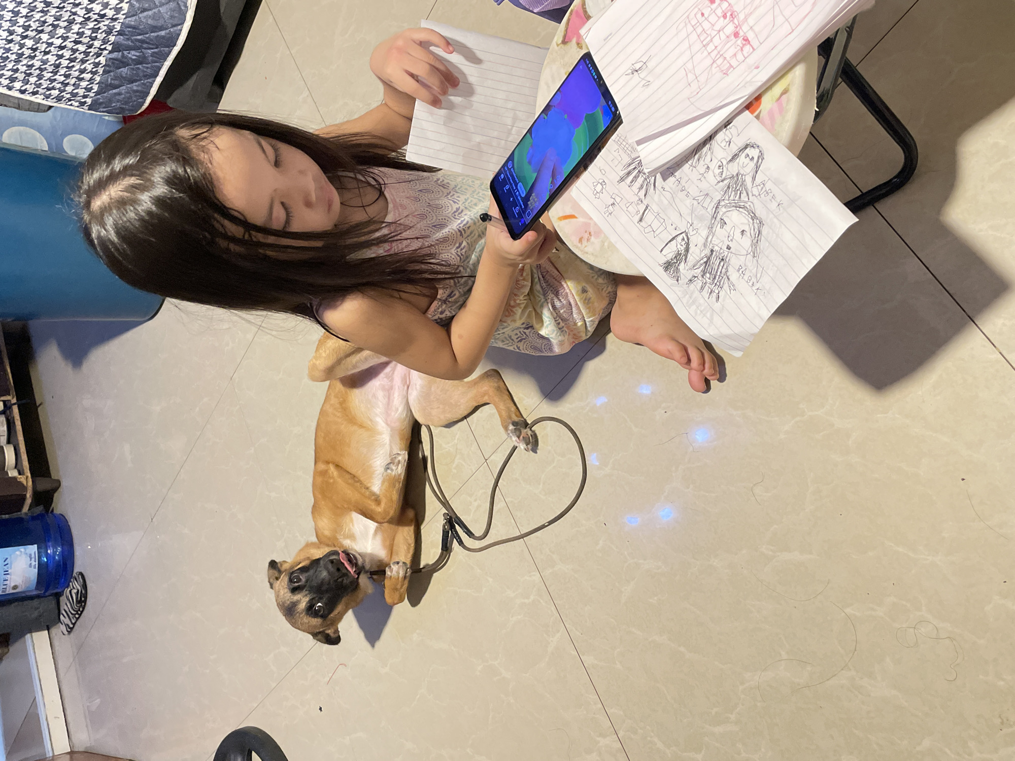 A young girl, with dark hair pulled back, is seated on the floor, engrossed in her smartphone. She wears a sleeveless, patterned top. Beside her, a tan-colored dog lies on its back, tongue out, with a leash loosely wrapped around its body. The floor is tiled and appears to be indoors, with a blue water dispenser, a blue trash bin, and a patterned blanket visible in the background. Scattered papers with drawings are also present near the girl. The scene suggests a casual, domestic setting, possibly a living room or a play area, within a residential dwelling in Davao City, Philippines.