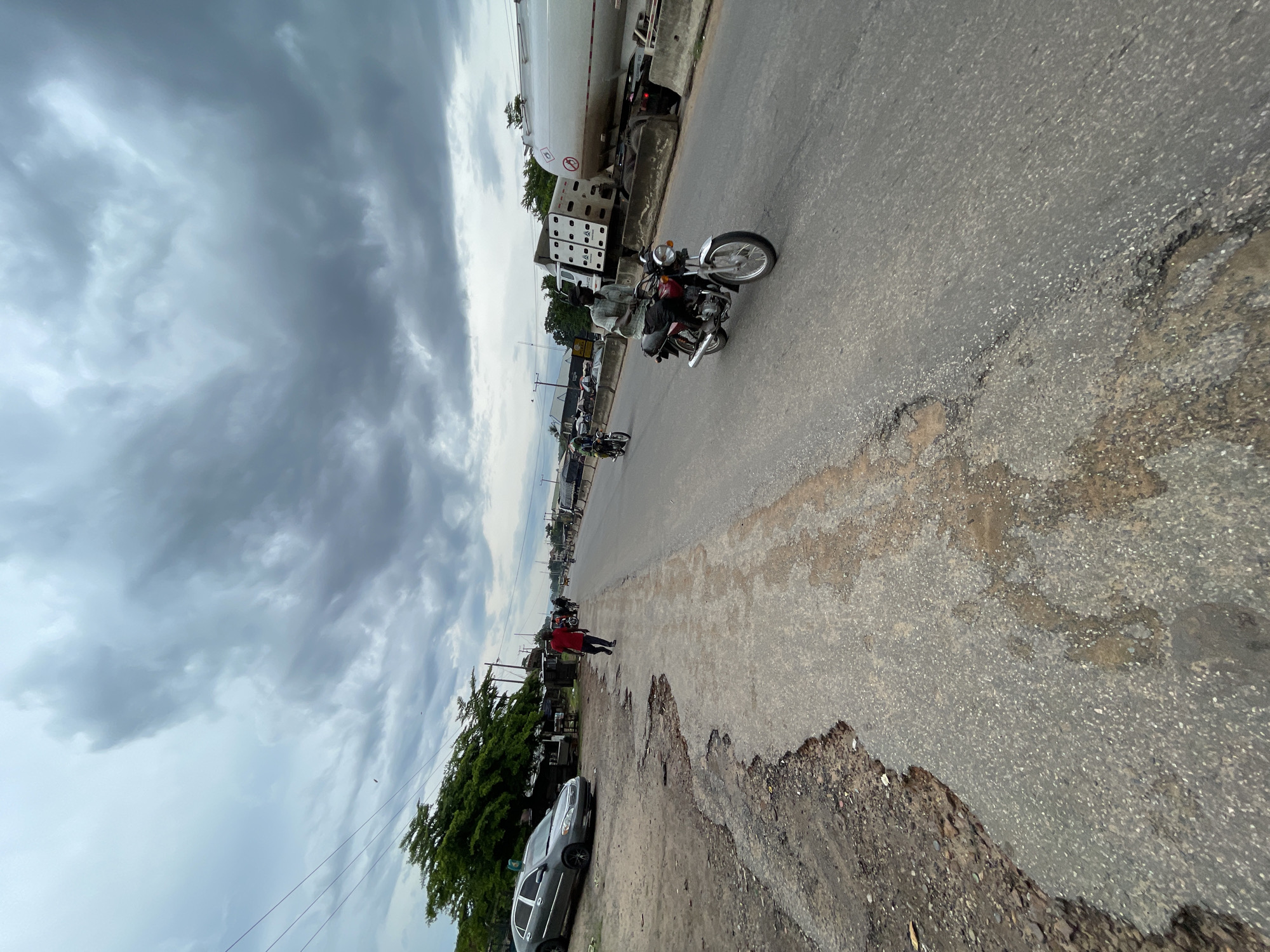 The image depicts a street scene in Lokoja, Nigeria, under a cloudy sky that suggests an overcast day, possibly before or after rain. The road surface is rough and shows significant wear and tear, with large potholes and eroded edges.
Several vehicles are present, including a large white tanker truck parked on the left side of the road. Further down the road, a motorcycle carrying two people is visible, with the rider wearing a helmet. Another motorcycle is seen further in the distance. A dark-colored car is parked on the side of the road, near some trees and small market stalls.
A person in a red shirt is walking along the side of the road, facing away from the camera. The overall impression is one of a busy, perhaps somewhat chaotic, urban or semi-urban environment. The lighting is diffused due to the cloud cover, casting a soft, even light on the scene. No discernible text is visible in the image.