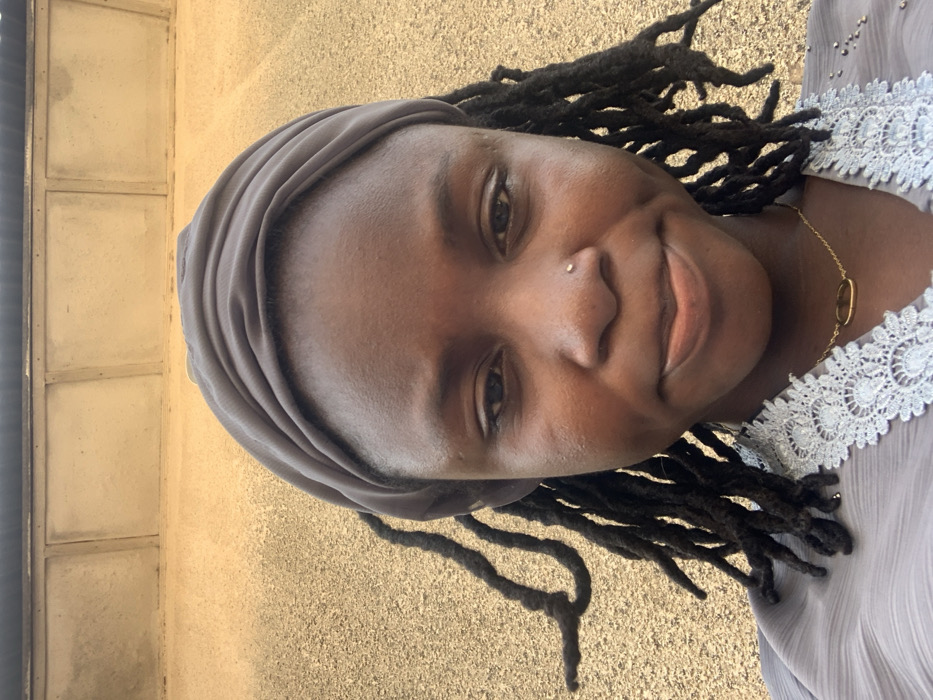 A close-up, chest-up photograph taken outdoors in Bojude, Nigeria, features a Black female individual. She wears a dark grey fabric headwrap and a light grey garment with white lace trim at the neckline. Dark dreadlocks are visible framing her face. A small, metallic stud is present in her left nostril, and a thin gold-colored necklace with an oval pendant is worn around her neck. Her eyes are open, and she displays a slight smile. The background consists of a textured, light brown-tan wall, with a darker horizontal band and three recessed rectangular panels visible in the upper portion, consistent with a building's facade. The scene is illuminated by diffused daylight.