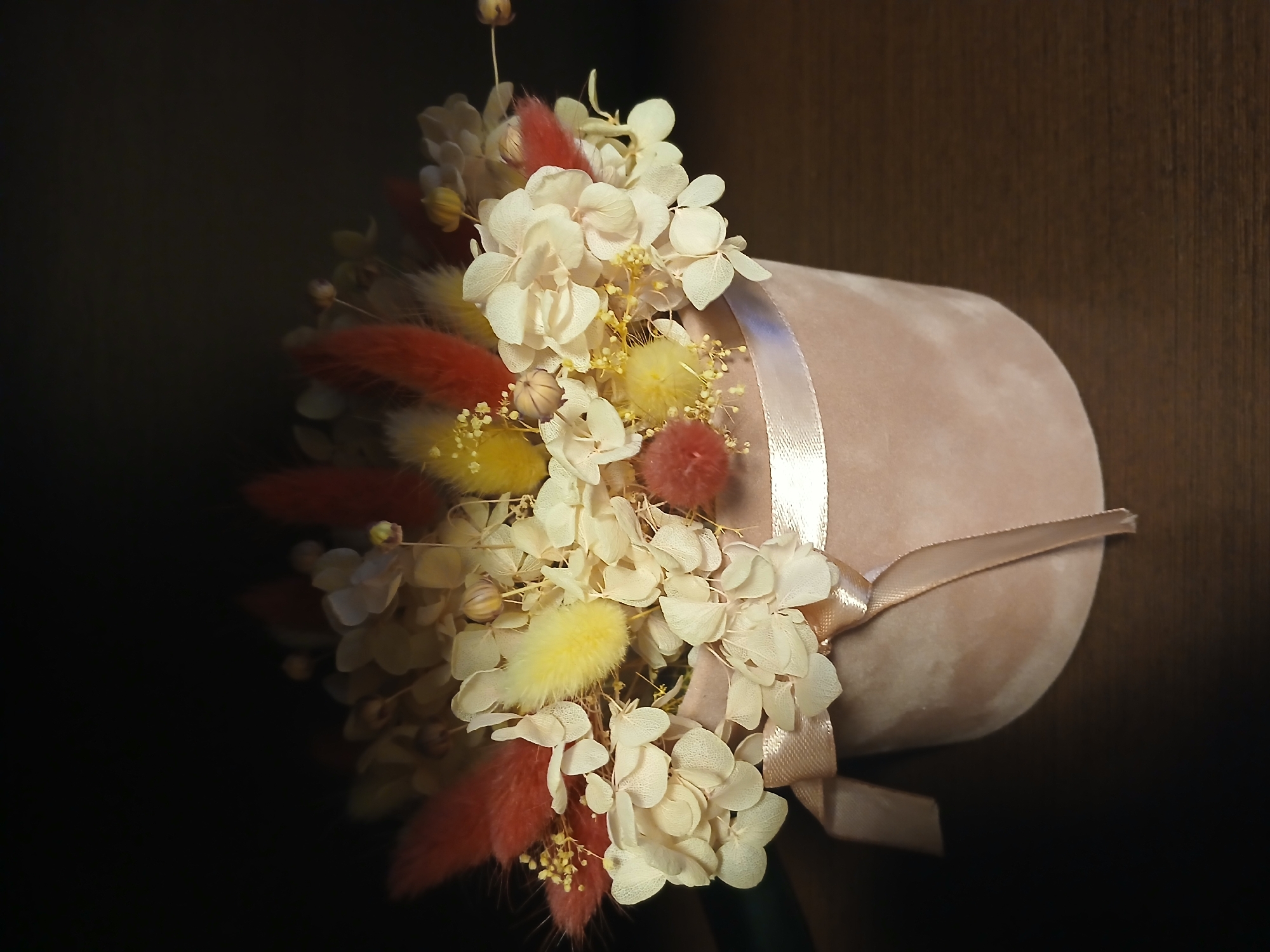 A studio shot depicts a floral arrangement in Kamianske, Ukraine. The arrangement features a cylindrical, pink velvet container tied with a satin ribbon. The container is filled with dried flowers including white hydrangea blossoms, red and yellow feathery stems, and small yellow pom-poms. The background is dark, emphasizing the light colors of the arrangement.