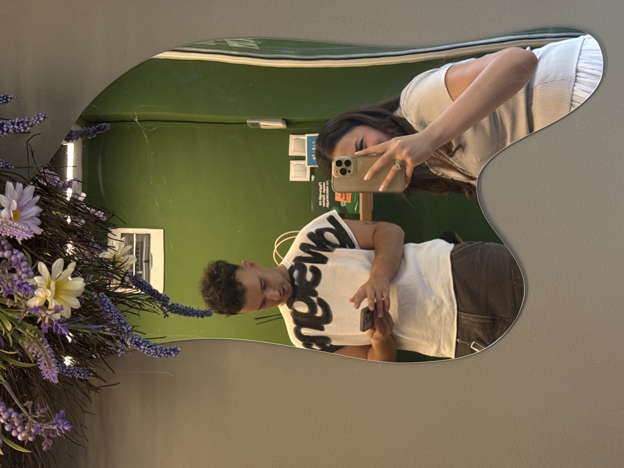 A couple is reflected in a mirror. A woman with long dark hair is holding a cell phone and taking a photograph. A man with curly dark hair is holding a cell phone. The mirror is hanging on a gray wall and is decorated with a bouquet of lavender and white flowers. The background is a green wall with a series of papers and signs posted on it. The location appears to be an interior space, potentially in Sofia, Bulgaria.