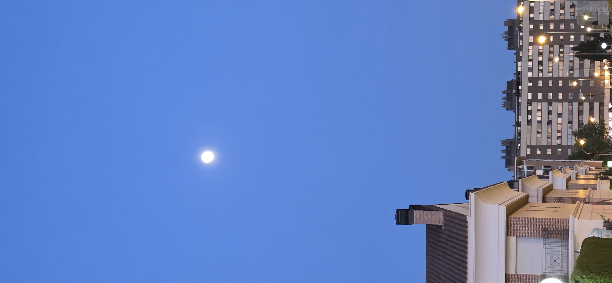 The image depicts a nighttime scene in Huesca, Spain, with a clear blue sky dominated by the moon. Below the sky, a tall, multi-story building with numerous illuminated windows is visible. A smaller structure with curved architectural details is also present, along with glimpses of street lighting. The image seems to be taken from a low angle.