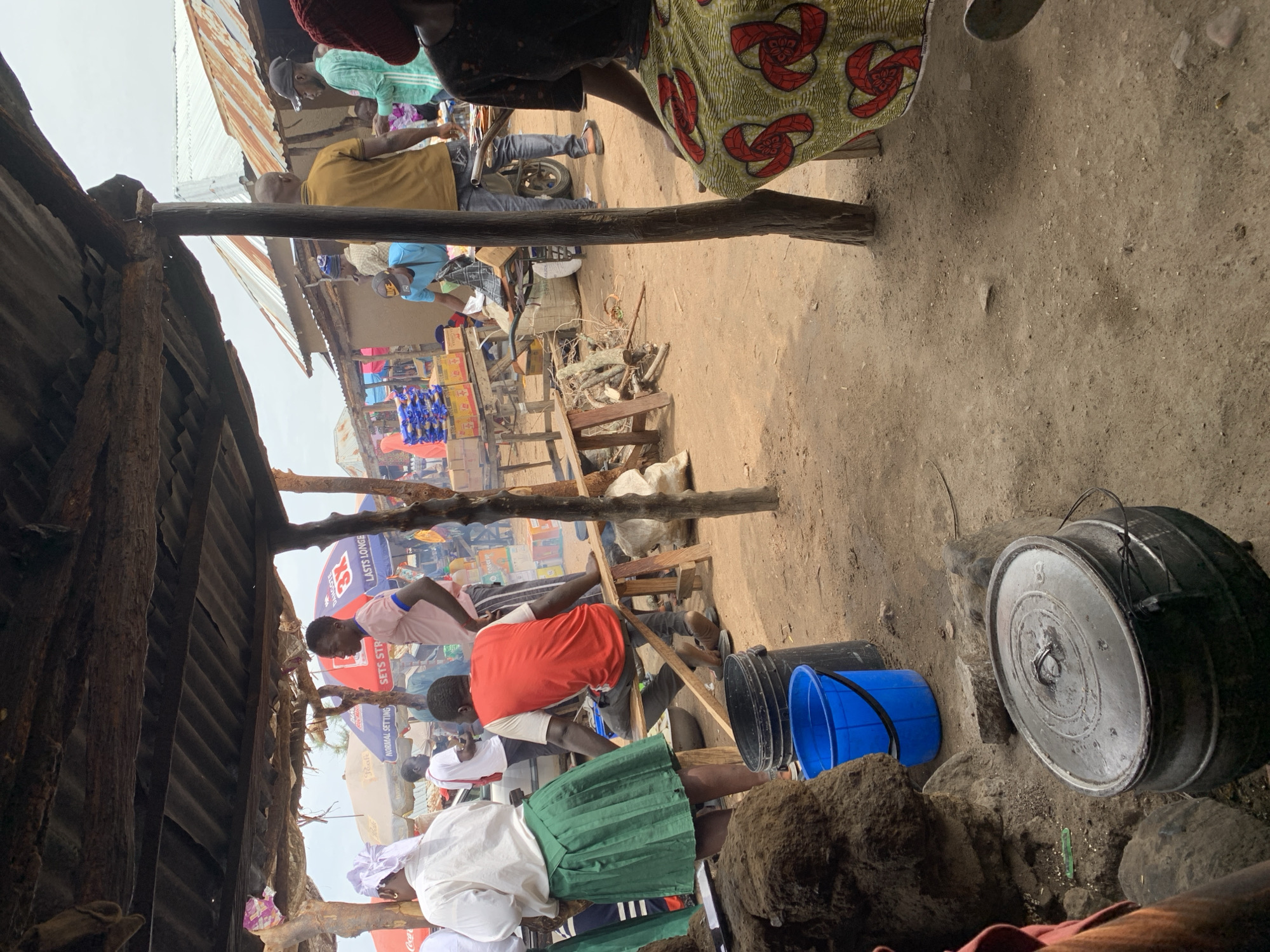 The image depicts an outdoor market scene in Dull A, Nigeria. Several people are present, some are standing, while others are seated. In the foreground, a large metal pot with a lid sits on the ground, possibly used for cooking. To the left, a blue plastic bucket and a black plastic container are visible. The environment appears to be dusty and rural, with makeshift structures providing shade from the sun. In the background, there are stalls with various goods displayed, including packaged food items. The overall atmosphere suggests a busy marketplace environment.