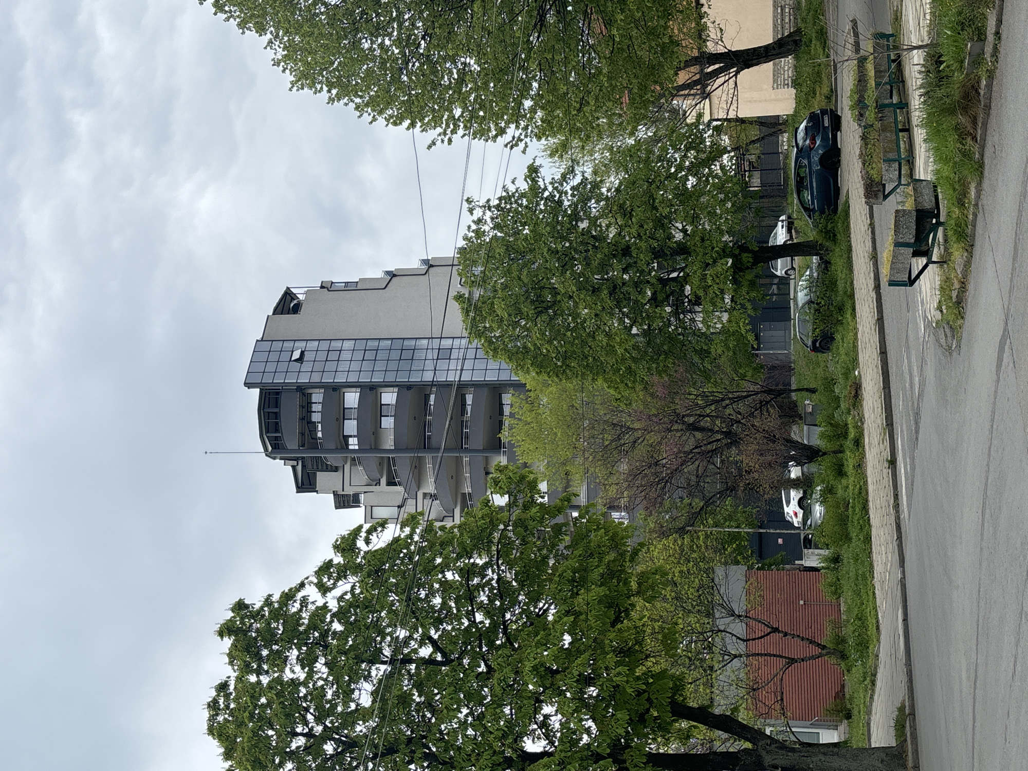 The image displays an urban or suburban street scene on an overcast day. A prominent, tall, modern building occupies the left side of the frame, characterized by numerous windows, extensive glass facades, and individual balconies. Its upper floors exhibit a stepped-back design, and a slender antenna or mast is visible on its roof.
In the foreground and midground, several trees with vibrant green leaves suggest a spring or summer setting, their branches extending across the scene and partially obscuring the sky and building. Utility lines are visible, stretching across the upper-mid portion of the image.
To the right, a paved road or street runs alongside a grassy verge. Several vehicles are parked on this grassy area, including a dark SUV and a white car. Two weathered park benches are also present on the verge. In the lower right corner, a smaller structure with a reddish-brown roof is partially visible. The sky is uniformly cloudy, indicating an overcast atmosphere. No human activity or discernible text is present in the scene.