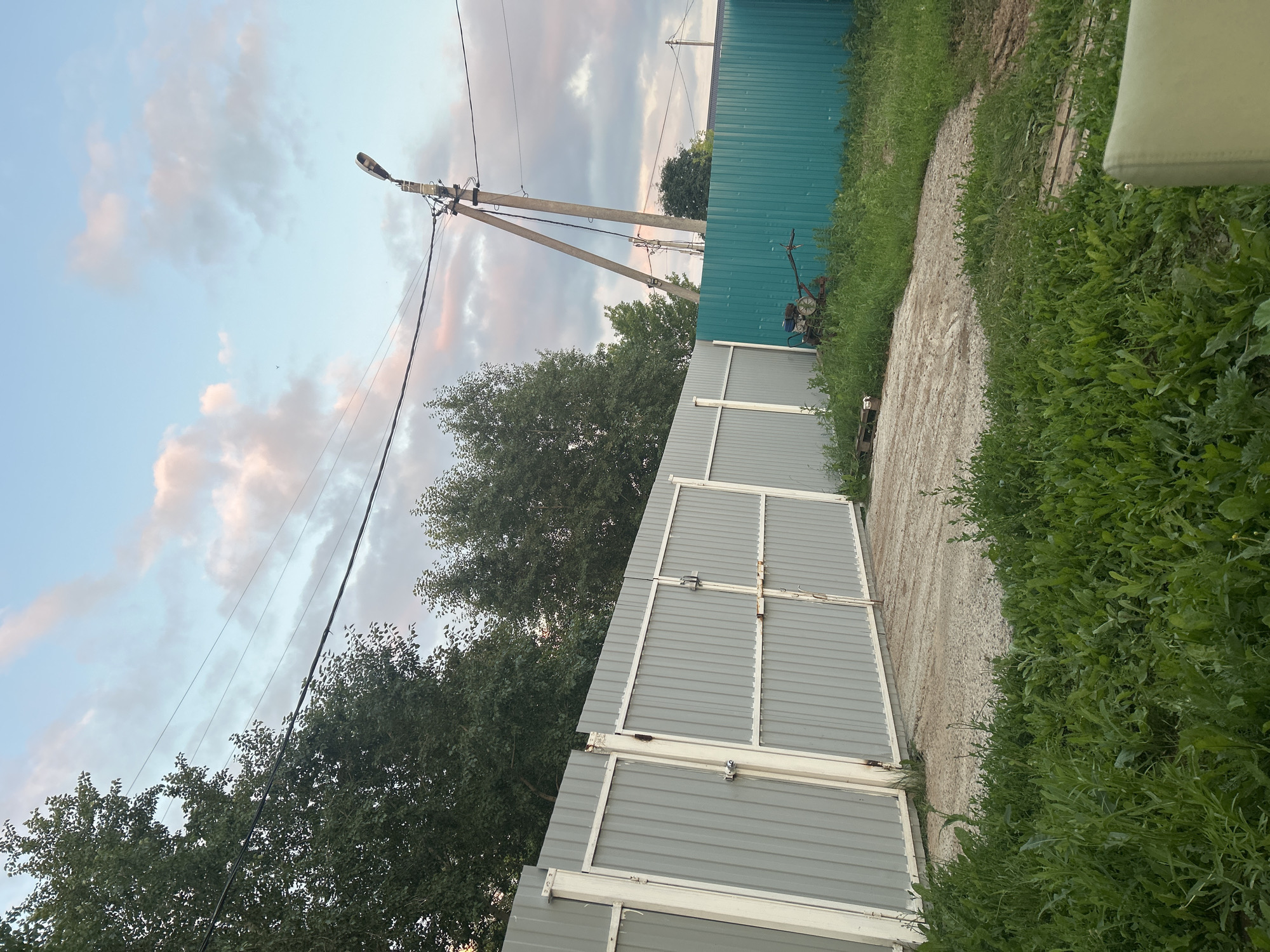 The image depicts an outdoor scene with a gray corrugated metal fence with white frames and two gates, one of which is slightly ajar. Adjacent to this fence is a teal corrugated metal fence. In the foreground, there is green grass and a gravel path leading to the gray fence. Behind the teal fence, a small red and blue tiller machine is visible in the grass. Power lines are strung across the sky, supported by utility poles. The sky is a blend of blue with pink and white clouds, suggesting either sunrise or sunset. The overall setting appears to be a rural or suburban area, possibly a backyard or a plot of land. There are no people visible in the image. The environment is natural with some man-made structures.