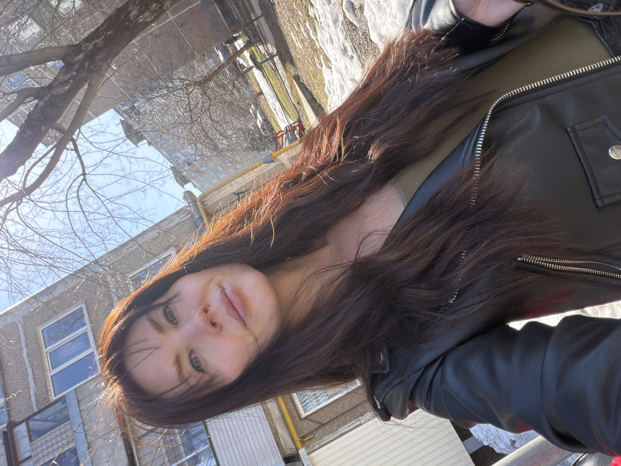 A young woman is taking a selfie outdoors in Perm, Russia. She has long, brown hair and is wearing a black leather jacket over a green top. The background shows a multi-story apartment building with windows, some bare trees, and patches of snow on the ground. The lighting suggests it is daytime.