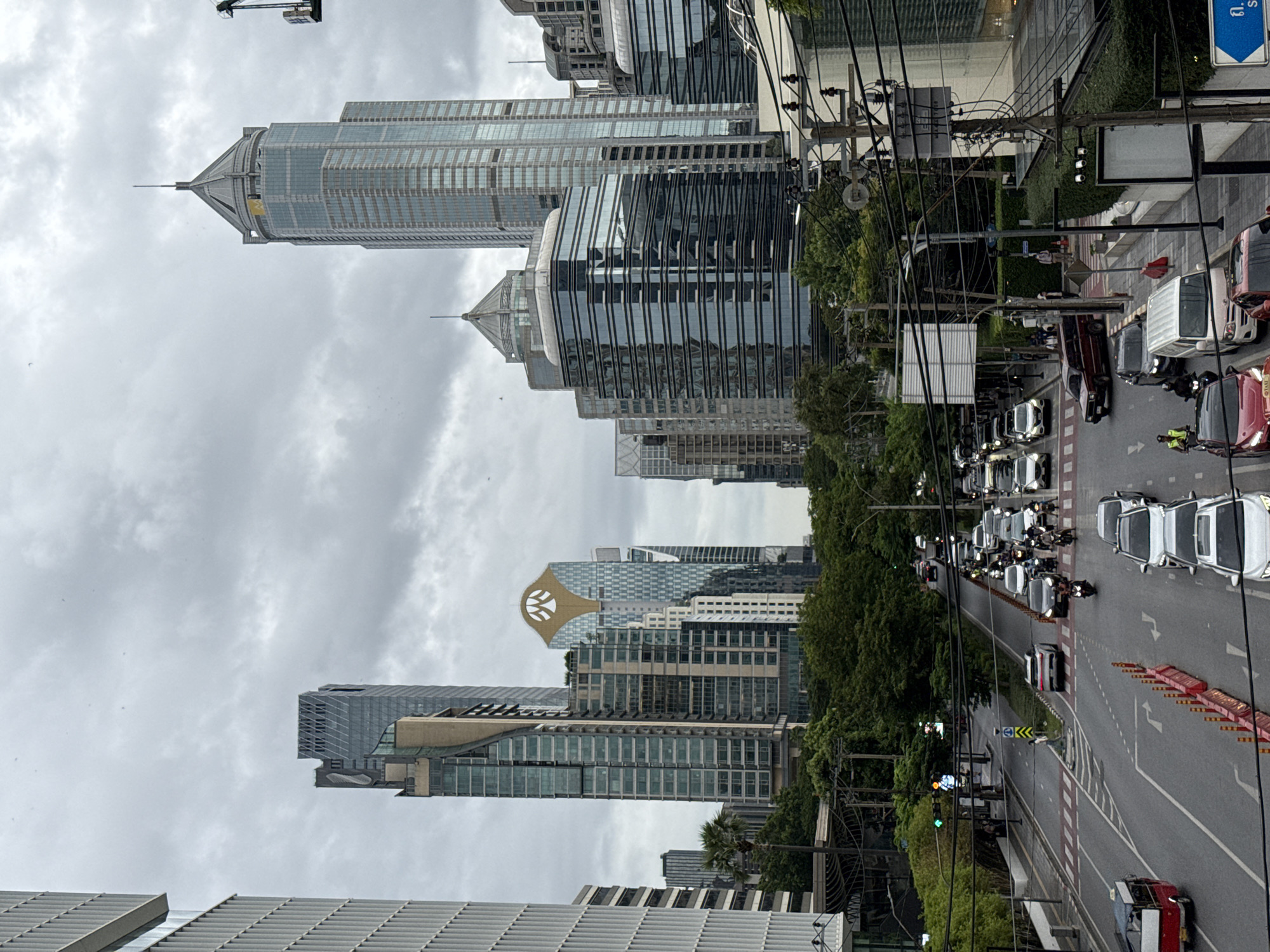 This is a street-level, medium-distance, over-the-shoulder view of a busy intersection in Bangkok, Thailand, on a cloudy day. The scene is dominated by modern, tall buildings on either side of a multi-lane road. In the foreground, vehicles are stalled in traffic, including a red and white tuk-tuk, several cars, and a number of motorcycles. Pedestrians are visible near a crosswalk. The background features a dense cluster of skyscrapers, characterized by glass facades and sharp architectural designs. Lush green trees are interspersed between the buildings, softening the urban landscape. The overall atmosphere is one of bustling city life under a heavy, overcast sky.