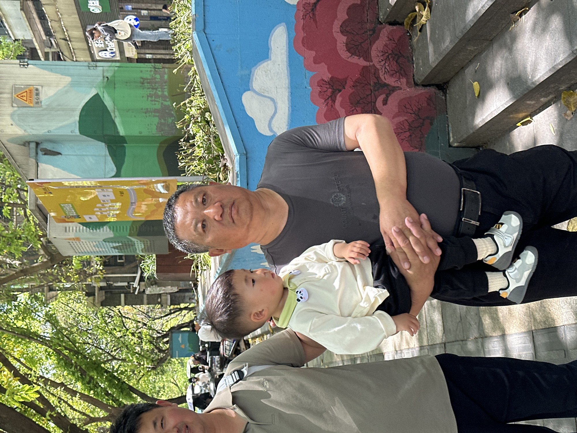 The image depicts two men holding a toddler in an outdoor setting. The men appear to be family members, possibly father and grandfather, holding the child between them. The child is wearing a light-colored shirt and dark pants with white shoes. The men are wearing casual clothing.
The background shows a cityscape with trees, buildings, and street art. The painting on the wall is red and pink. The location appears to be in an urban area with pedestrian traffic. The picture indicates the location is in Shapingba District, China.