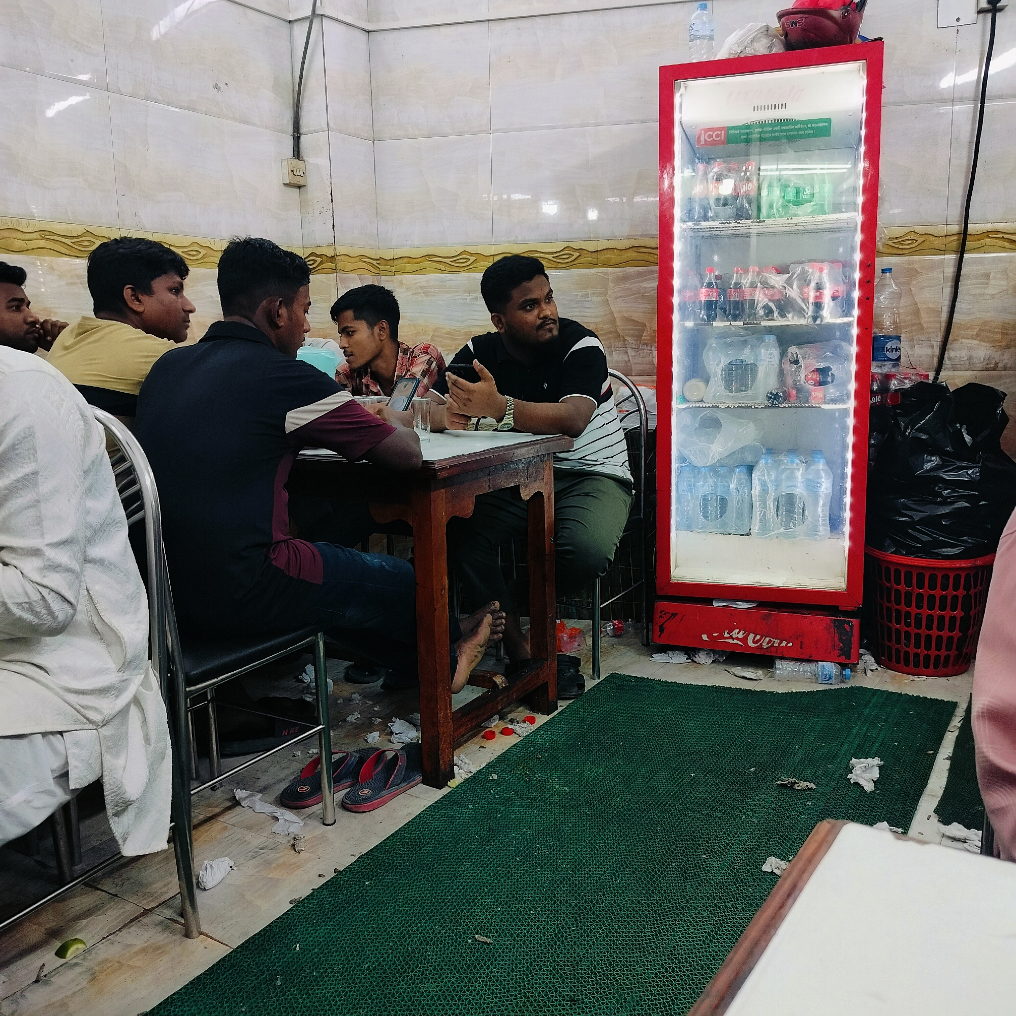 In a dimly lit, utilitarian indoor setting resembling a small eatery or tea stall in Dhaka, Bangladesh, a group of young men are seated around a wooden table. The walls are tiled with a beige and brown pattern, and a red Coca-Cola branded refrigerator stands prominently to the right, stocked with beverages. The floor is partially covered by a green textured mat and scattered with used tissues and some discarded plastic bottles, suggesting a casual and possibly frequented establishment.
Four men are clearly visible. On the far left, a man wearing a white garment sits with his back to the camera. Next to him, a young man with short black hair in a yellow t-shirt is seen from the side, looking to his left. To his right, another man with short black hair, wearing a dark t-shirt with white and maroon stripes, is holding a mobile phone and looking upwards, with a watch on his left wrist. Across from him, a man in a plaid shirt is partially visible, looking towards the right.
On the table, glasses and possibly a phone are present, indicating consumption and activity. The overall atmosphere appears informal, with the men engaged in conversation or their own activities. The presence of the refrigerator and the general decor suggest a local food and beverage outlet.