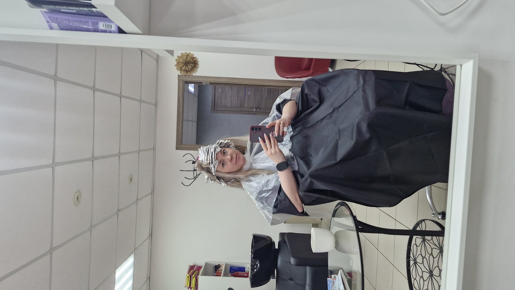 A woman with blonde hair wrapped in foil is taking a selfie in a salon mirror, wearing a black cape. She holds a purple phone and wears a black smartwatch. The salon setting includes a hair dryer, shelves with products, a door with a golden ornament, and a red chair. The location is likely in Otchor, Russia.