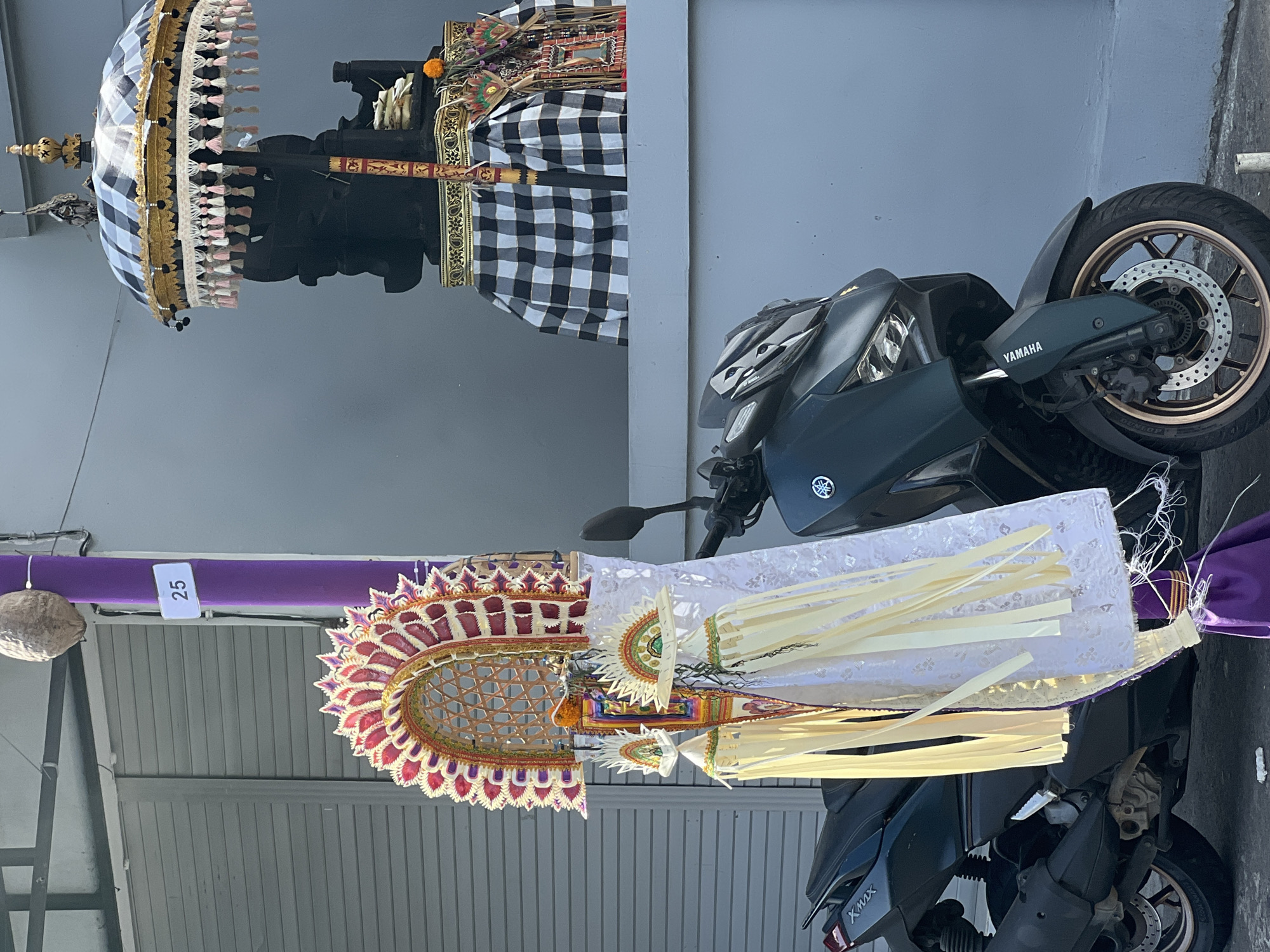 In front of a grey building, a dark green Yamaha XMAX motorcycle is parked. Attached to a purple pole to the left of the motorcycle are several traditional Balinese decorations. These include a large fan-shaped ornament at the top, a white lace-like fabric with yellow streamers hanging down, and a smaller fan-shaped ornament below the larger one. Behind the motorcycle and to the right, a dark statue, likely of a deity, is adorned with a black and white checkered cloth and a black and white checkered umbrella with gold trim and pink and white tassels. A small orange flower is visible on the statue's base. A white square with the number '25' is attached to the purple pole. The scene appears to be outdoors in an urban environment.