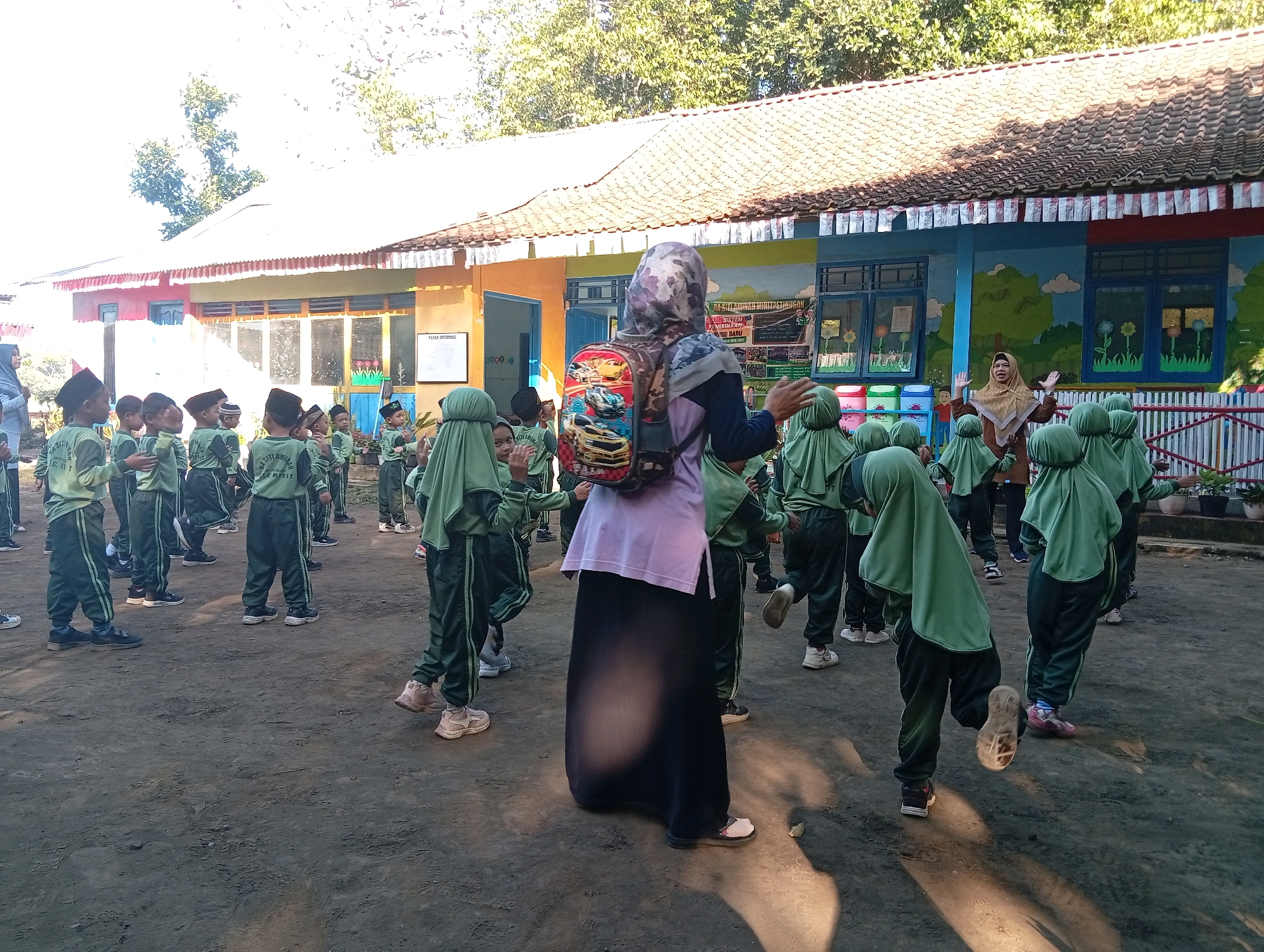 A group of young children wearing green uniforms and headscarves are gathered in an outdoor area, likely a schoolyard, in Miritpetikusan, Indonesia. They appear to be participating in an activity, possibly a lesson or exercise, led by a teacher or adult who is also present. The adult, wearing a patterned headscarf and a long dark skirt, stands in the foreground facing away from the camera, with a backpack on. The children are arranged in rows and some are looking towards the adult. The background features a building with a tiled roof and colorful murals on its walls, typical of a school environment. The ground is unpaved, and shadows indicate a bright sunny day. There are also some potted plants visible.