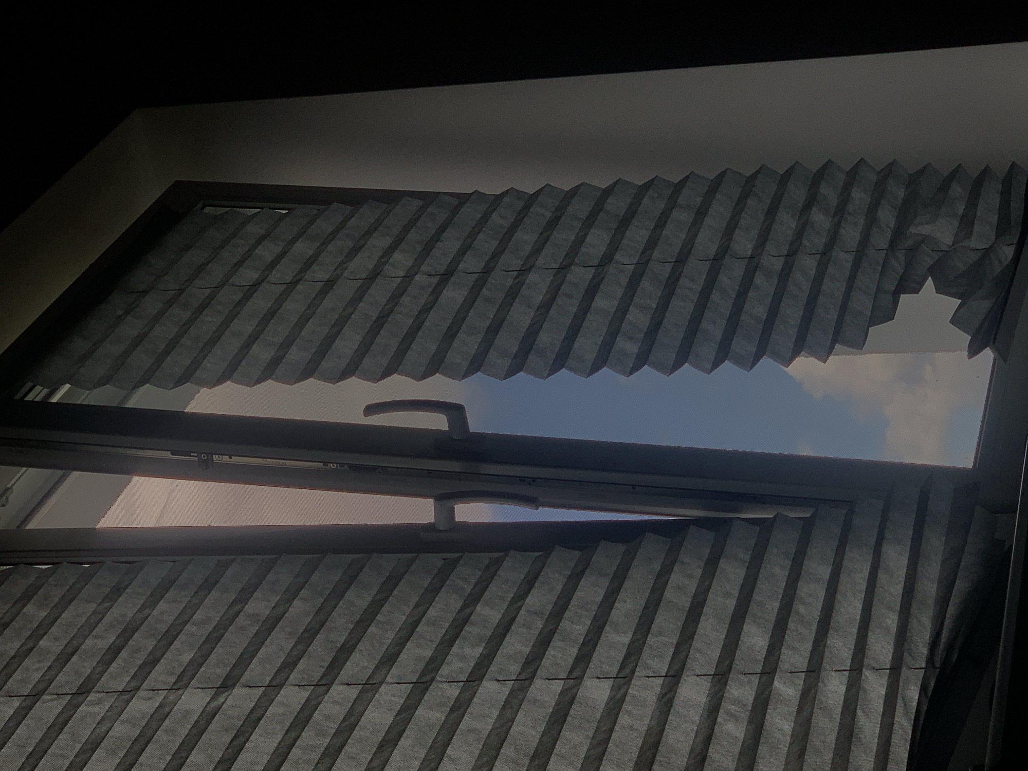 The image shows an interior view of a partially opened window with gray pleated blinds. The blinds are pulled to the sides, revealing a sky with clouds. The window frame is dark, and the surrounding wall is light-colored, transitioning to dark on the right side of the frame. The scene is consistent with a typical window in a residential or office building. There is no information in the image to suggest a specific location in Minsk, Belarus, or any details about people, actions, or interactions.