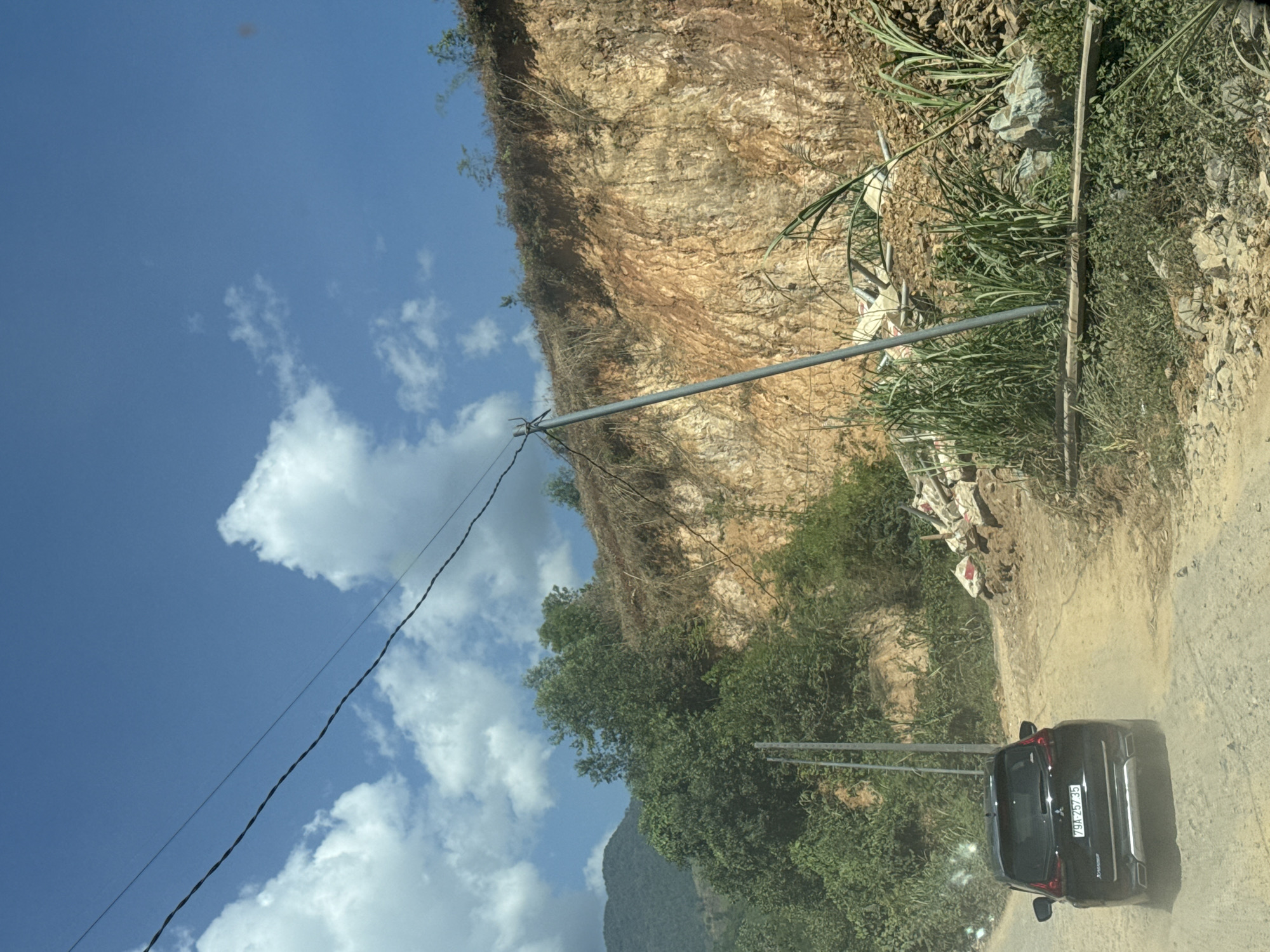A black SUV with license plate "79A-257.35" is driving on a dirt road in a rural area, possibly in or near Nam Khánh Vĩnh, Vietnam. The road is dusty, indicating the SUV is in motion. To the right of the road, a steep, eroded cliff face is visible, composed of reddish-brown soil and rock. Vegetation, including tall grasses and some trees, grows on and around the cliff. A gray utility pole stands near the road, supporting power lines that stretch across the sky. In the background, a mountain covered in greenery can be seen. The sky is bright blue with scattered white clouds. Debris, including what appear to be construction materials like cement bags and pipes, is piled near the base of the cliff.