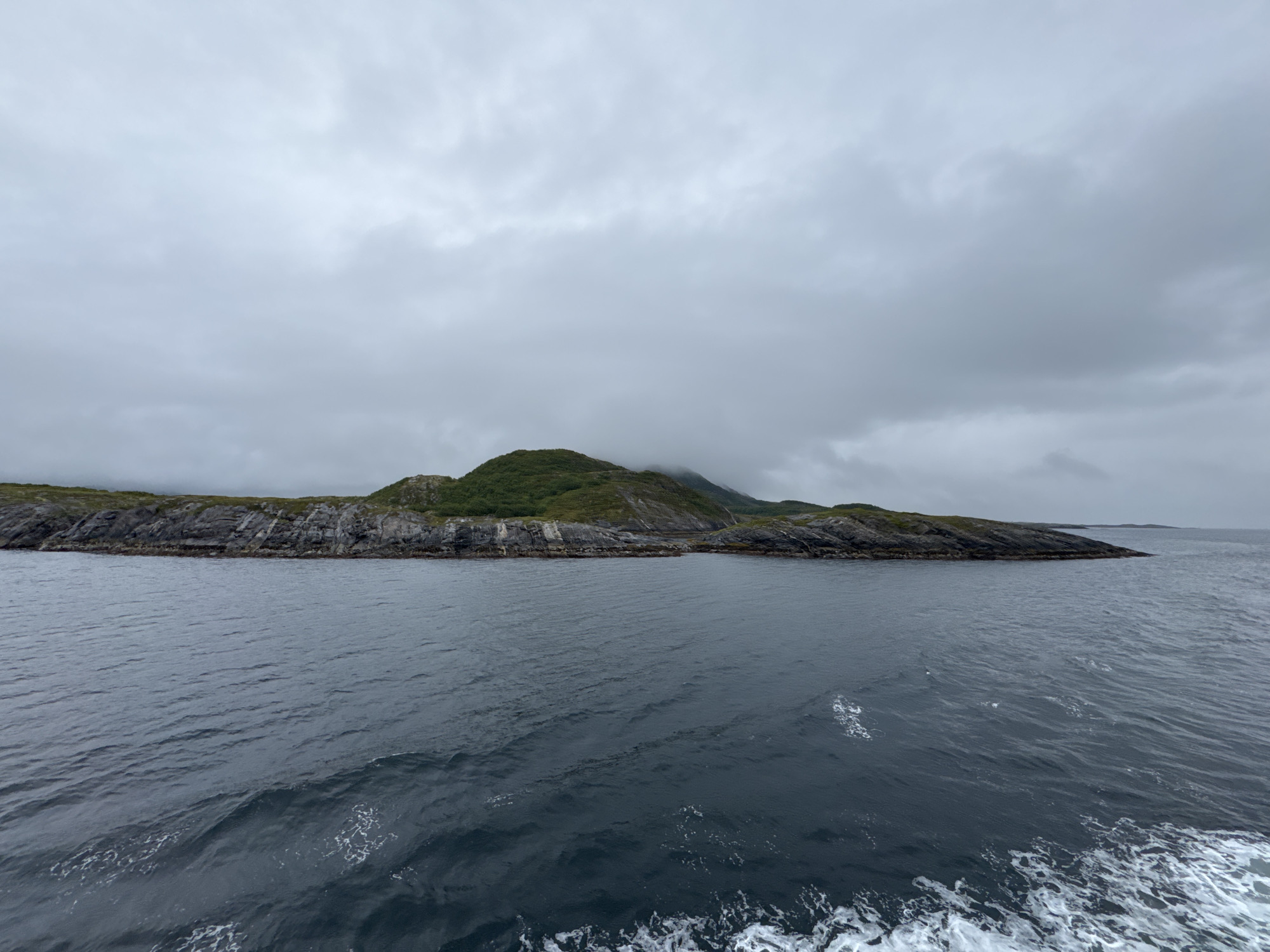 An overcast, grey sky looms above a body of dark, choppy water. In the middle ground, a long, narrow island stretches horizontally across the frame. The island's foreground is characterized by rugged, grey rock formations, interspersed with patches of green vegetation. The island's interior rises into gently sloping hills covered in lush green foliage, with some sections obscured by mist. The water in the foreground exhibits white foam and ripples, suggesting movement. The overall scene depicts a coastal landscape under a cloudy sky in Demmahågen, Norway.