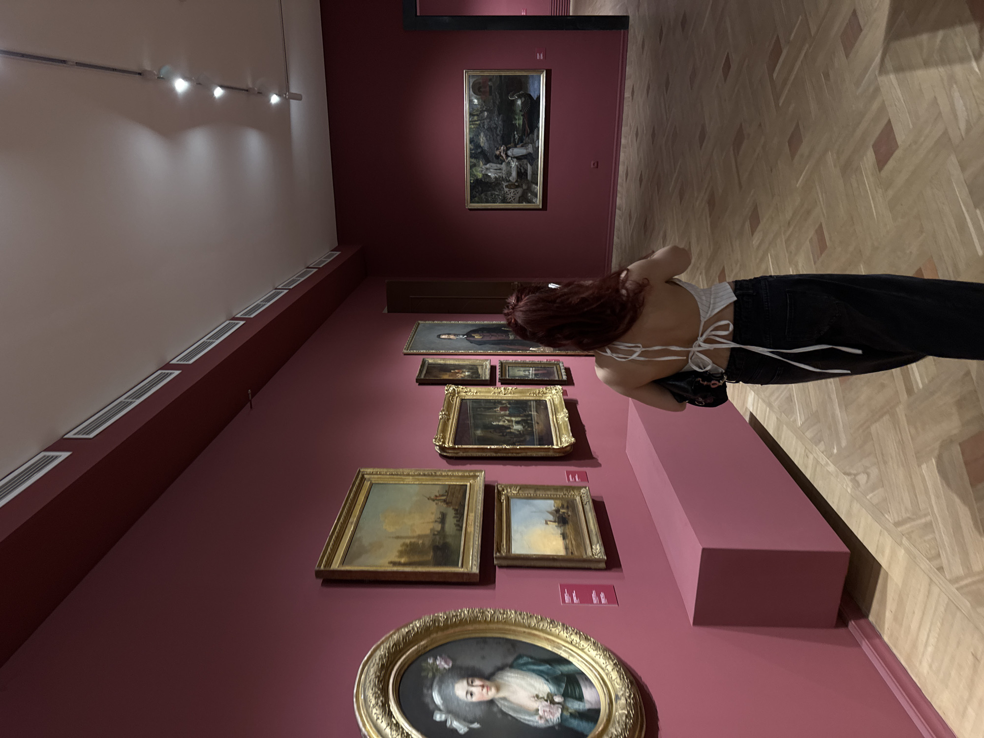 A person with long, auburn hair is standing in a museum, facing away from the camera, and looking at a painting on a deep red wall. The person is wearing a white halter top and dark jeans. The museum has a polished wooden floor and is lit by track lighting. There are several framed paintings on the wall. To the left, a large oval portrait of a woman in 18th-century attire is prominent. To the right, a rectangular landscape painting is visible. The location is identified as the National Gallery of Armenia in Yerevan, Armenia.