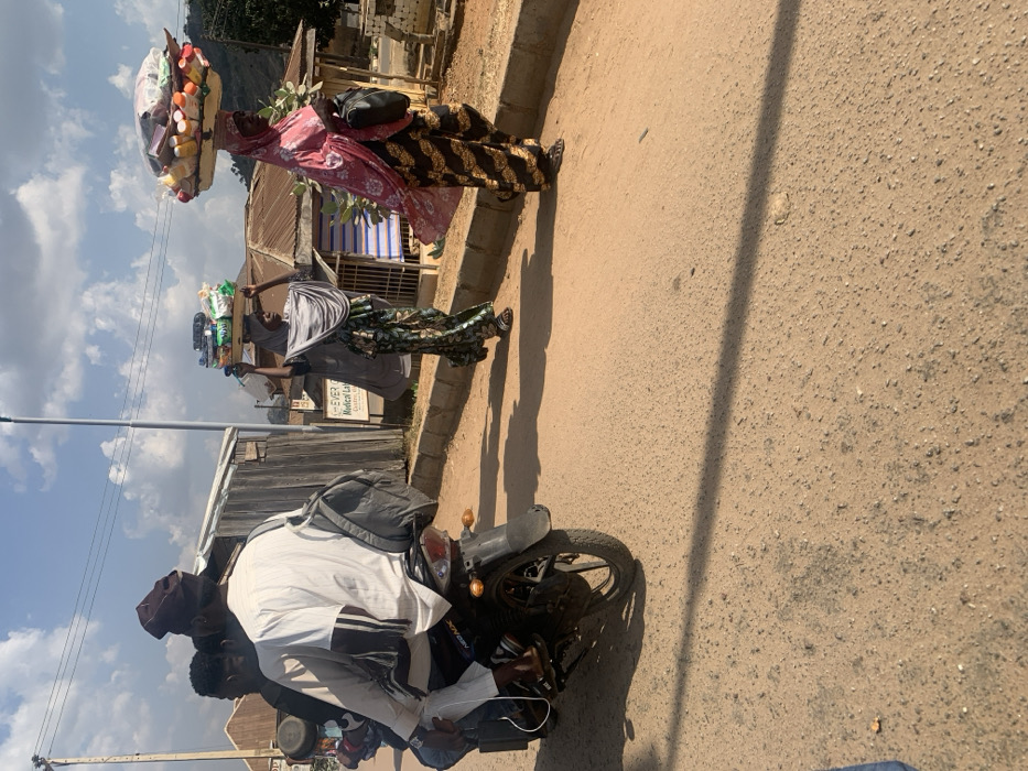The scene depicts a daytime street in Okengwe, Nigeria. In the foreground on the left, a black motorcycle carries two males, observed from the rear. The operator wears a white long-sleeved shirt, light trousers, and a dark cap, with a grey duffel bag slung over his back. A second male is positioned as a passenger directly behind him. On the right side of the frame, two women walk parallel to the road on an unpaved shoulder, which is separated from the main thoroughfare by a low concrete curb. The first woman, closer to the motorcycle, wears a patterned full-length dress and a grey headscarf, balancing a rectangular tray of packaged items on her head, which she supports with both hands. The second woman, positioned further to the right, wears a distinct patterned full-length dress and a pink headscarf. She carries a larger, mounded display of various packaged consumer goods on her head without manual support, and a black shoulder bag is slung on her left side. The road surface consists of worn asphalt and packed earth. The background features various single-story structures with corrugated metal roofs, including one with a partially visible sign reading "EVER Medical La...". Sparse vegetation is present, along with a prominent grey utility pole. Distant hills are visible on the horizon under a partly cloudy sky. Distinct, elongated shadows are cast across the road, indicating a strong light source from the left.