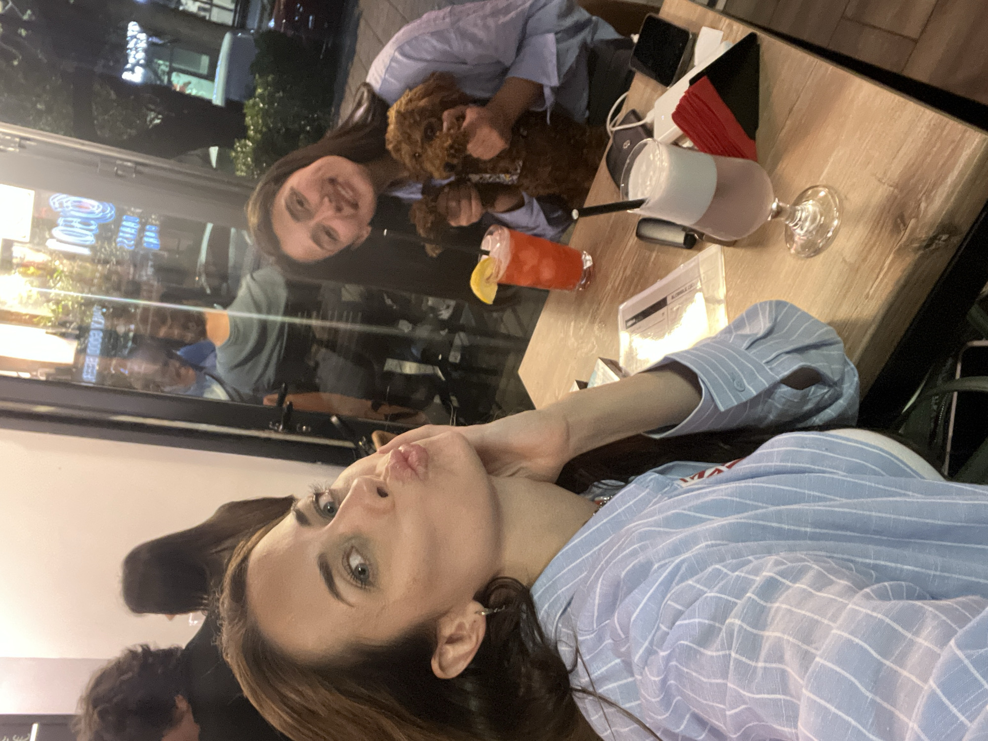 The image shows two women and a dog at an outdoor restaurant. The woman in the foreground is taking a selfie and is wearing a blue and white striped shirt. The other woman is holding a brown dog with a patterned collar. There are two cocktails on the wooden table, one is pink and one is orange with a lemon slice. In the background is a window showing a reflection of people and blue neon lights, possibly located in Bar, Montenegro. Other items on the table include a phone, a black wallet, a white power bank, and a container of red napkins.