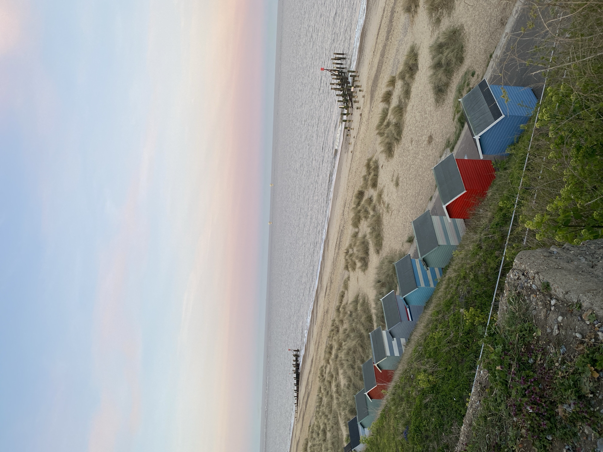 The photograph depicts a beach scene in East Suffolk, United Kingdom. Several rows of colorful beach huts line the right side of the frame, nestled among grassy dunes. A concrete embankment sits adjacent to the huts. The beach stretches out towards the sea, with a series of wooden groynes extending into the water. The sky is overcast with pastel hues, and the sea is calm. There are no people visible in the image.