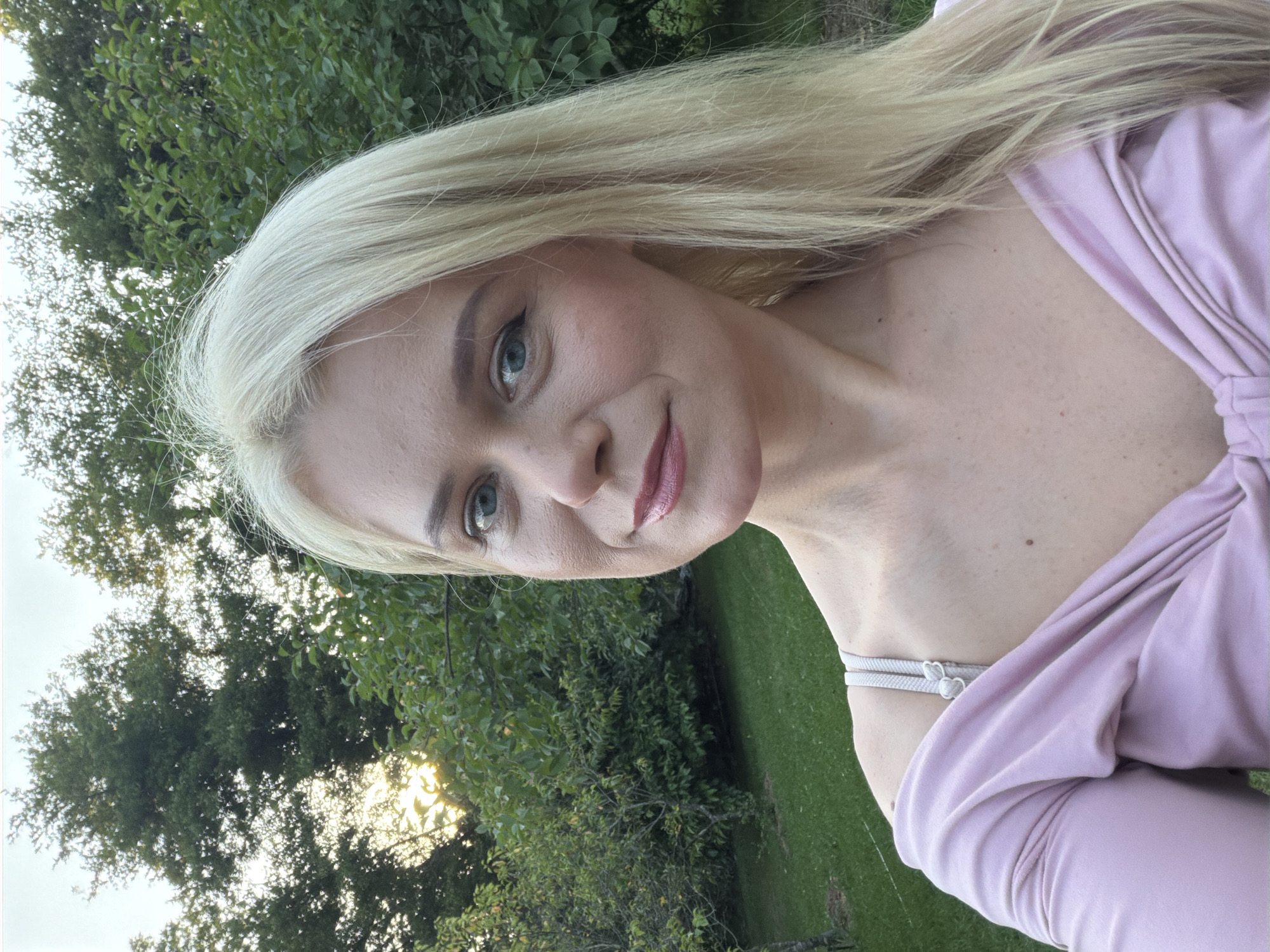 The media file is a portrait of a blonde woman with blue eyes, smiling at the camera. She is wearing a light pink off-the-shoulder top. The background is a natural setting with lush green trees and grass, suggesting an outdoor environment. The sun is visible through the trees, indicating it is daytime, likely late afternoon or early evening due to the warm lighting. The woman's makeup is subtle, with emphasis on her eyes and lips. The overall mood appears relaxed and pleasant. The location context provided is Sochi, Russia. There is no visible text in the image.