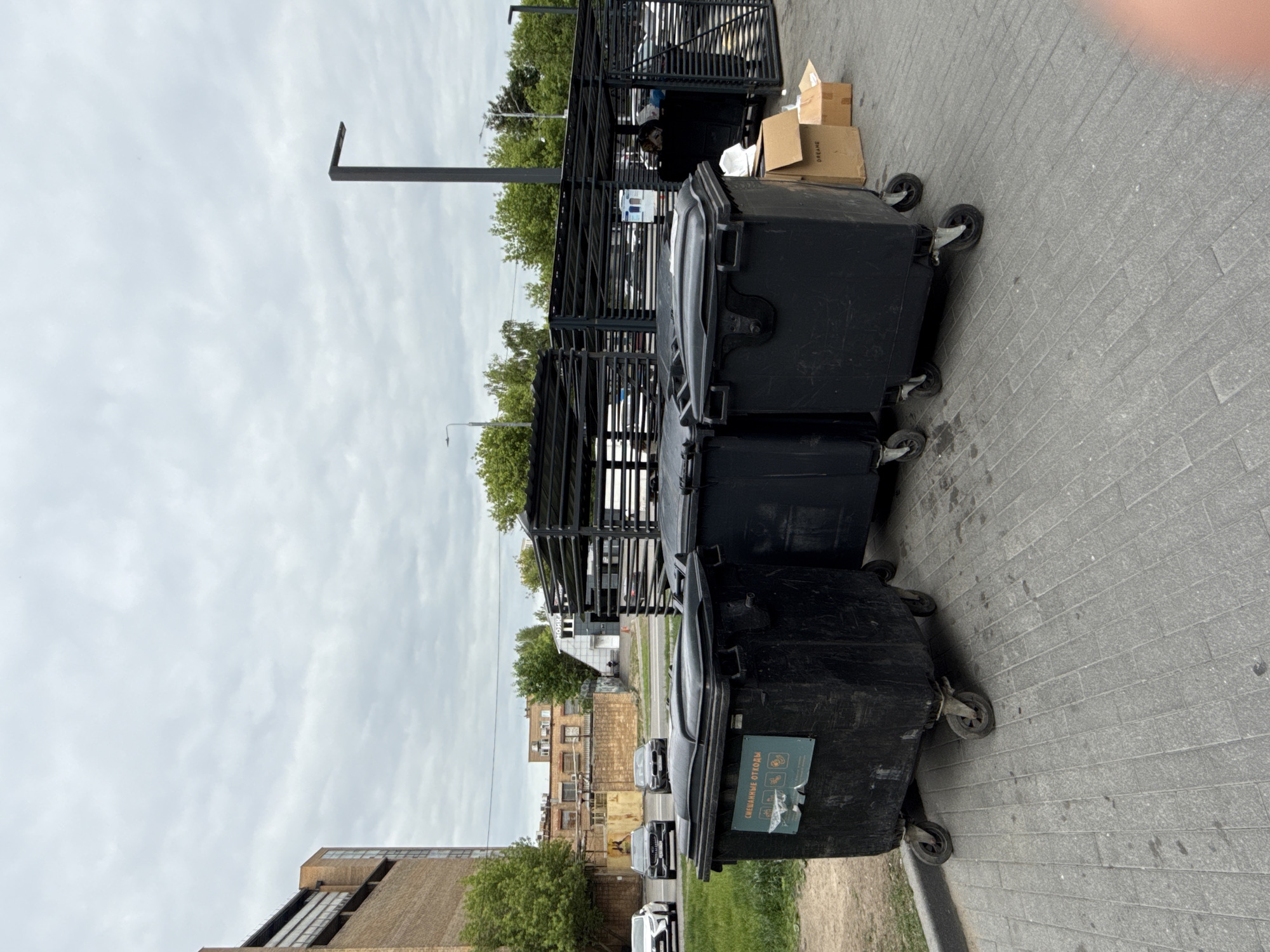 The image displays an urban scene in Moscow, Russia, primarily focused on a row of large, black, wheeled trash bins positioned on a grey paved surface, likely a sidewalk or alleyway. The sky is overcast with soft, light clouds, indicating daytime.
Adjacent to the bins, a substantial black metal structure, possibly a waste enclosure or bicycle storage unit, is visible. Beyond this structure, the background reveals a street with several cars and a collection of buildings. These buildings vary in style, including an older brick structure and another with a more modern design featuring rooftop windows or skylights. Green trees are interspersed throughout the urban landscape, adding patches of foliage.
A prominent detail is a sticker on the closest bin, displaying Cyrillic text that translates to "СМЕШАННЫЕ ОТХОДЫ" (Mixed Waste), accompanied by icons for different types of refuse, such as bottles, cans, and general waste, emphasizing its function for waste collection. Nearby, several brown cardboard boxes, one with the word "DREAME" visible, are placed on the ground. No individuals are present, and no active events are unfolding; the scene depicts a static, functional area dedicated to urban waste management.
