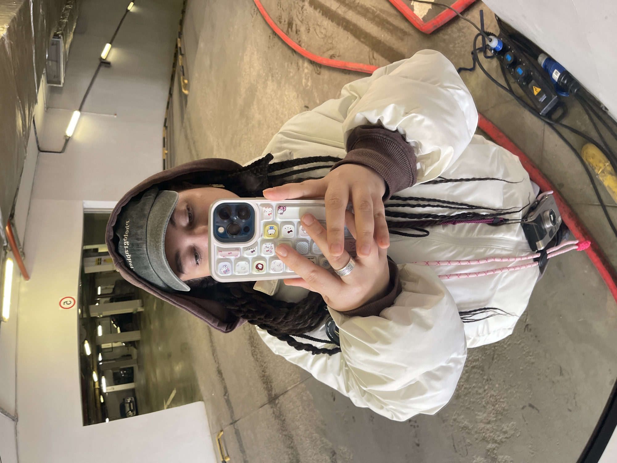 A person is taking a selfie in a mirror. The person is wearing a brown hooded sweatshirt, a white puffer jacket with black and white striped accents, and a baseball cap. The phone they are using has a white case with cartoon designs. The person is wearing a ring on their left ring finger. The reflection in the mirror shows a parking garage with visible lighting fixtures on the ceiling and cars parked in the distance. The location is Unknown, Unknown. Additional equipment like red cable guards and electrical distribution boxes are visible.