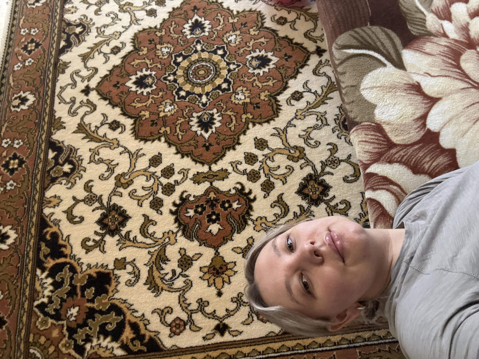 An indoor scene depicts a woman photographed from the upper chest level upwards, looking directly at the camera. She has light-colored hair and is wearing a light grey short-sleeved top. A small silver hoop earring is visible in her left ear. She exhibits a neutral to slightly smiling expression. Behind her, covering the wall, is a large ornamental rug with a cream background and intricate traditional patterns in shades of brown, black, and gold. To her right and in the lower foreground, a section of a sofa or cushioned furniture is visible, featuring a distinct, larger floral pattern in brown, cream, and reddish-brown tones. The image captures an individual within a residential interior. The location is the city of Chaikovsky, Russia.