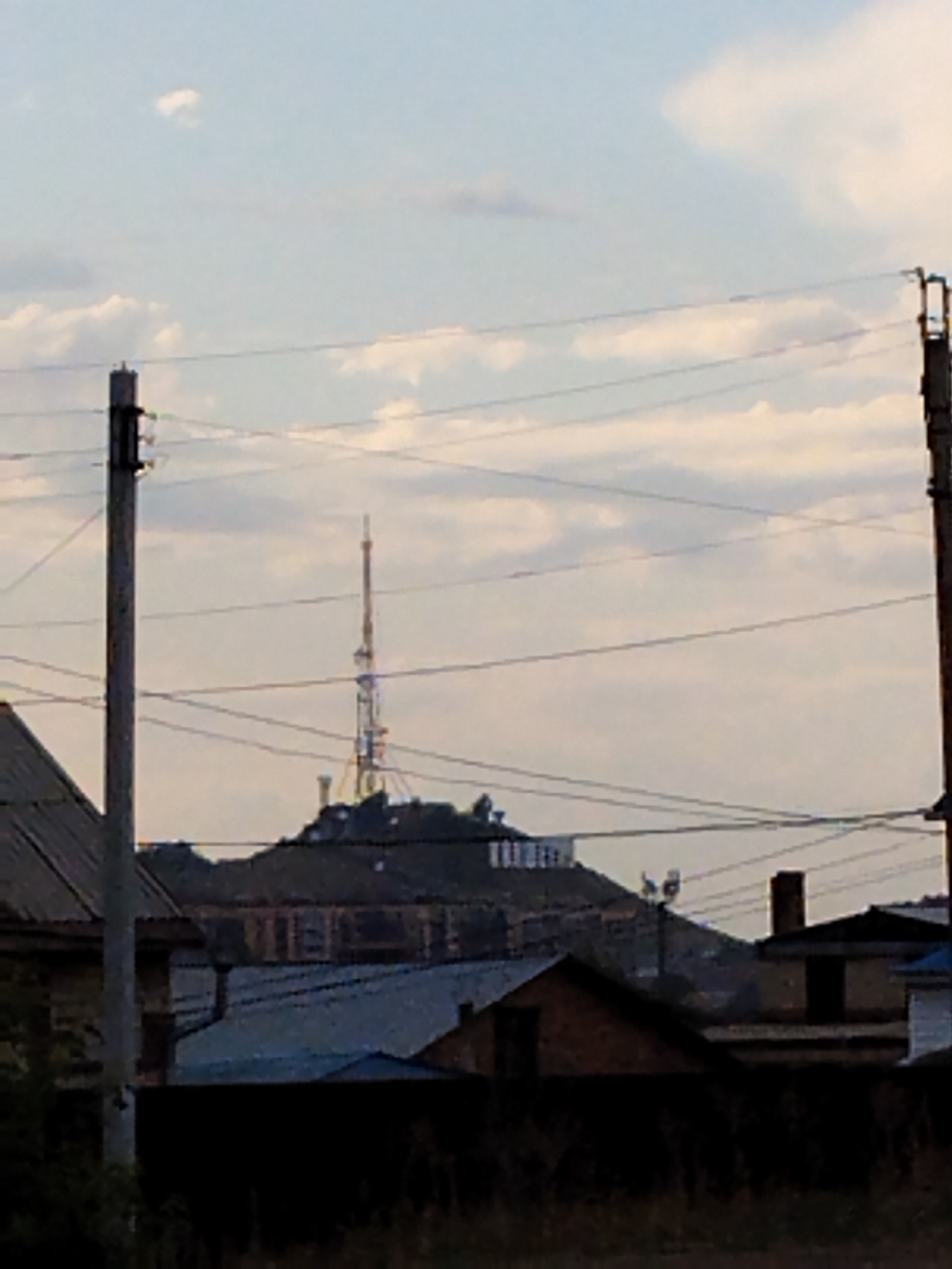 This image depicts a view of a hill in Kokshetau, Kazakhstan, with a prominent television tower and several buildings in the foreground. The sky is overcast with scattered clouds. The television tower is tall and slender, with a lattice structure. Below the tower, a town or village is visible, with rooftops and some structures clustered together. The foreground shows a variety of buildings, including houses with pitched roofs and some simpler structures. Power lines are strung across the scene, supported by utility poles. The overall impression is one of a semi-urban or rural landscape in Kokshetau.