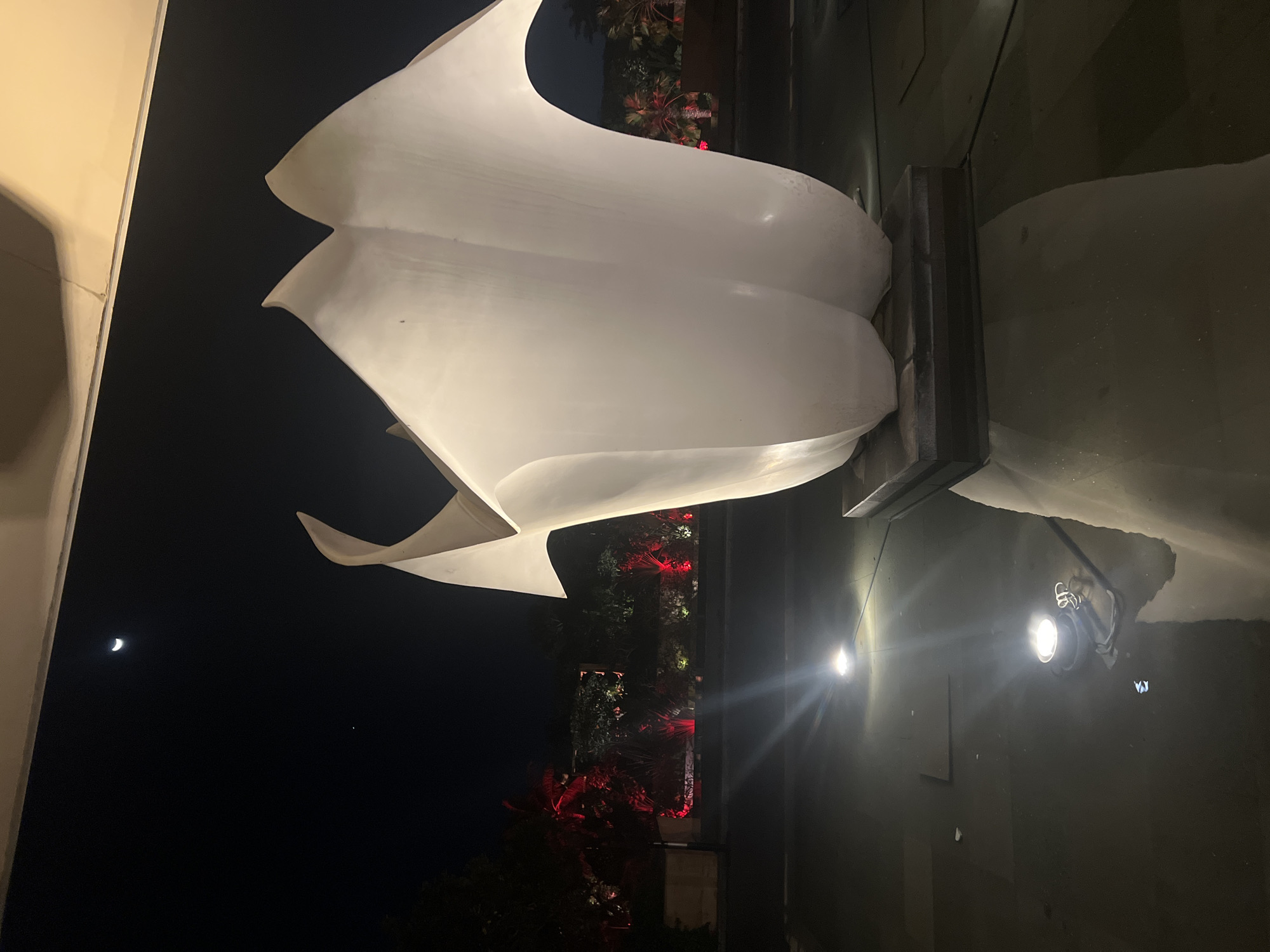 At night, a large white sculpture rests on a dark pedestal surrounded by water. The sculpture is illuminated by nearby lights. In the background, trees and foliage are visible, lit by red lights. A crescent moon is visible in the dark sky. The location is in the city of Kuta Selatan, Indonesia.