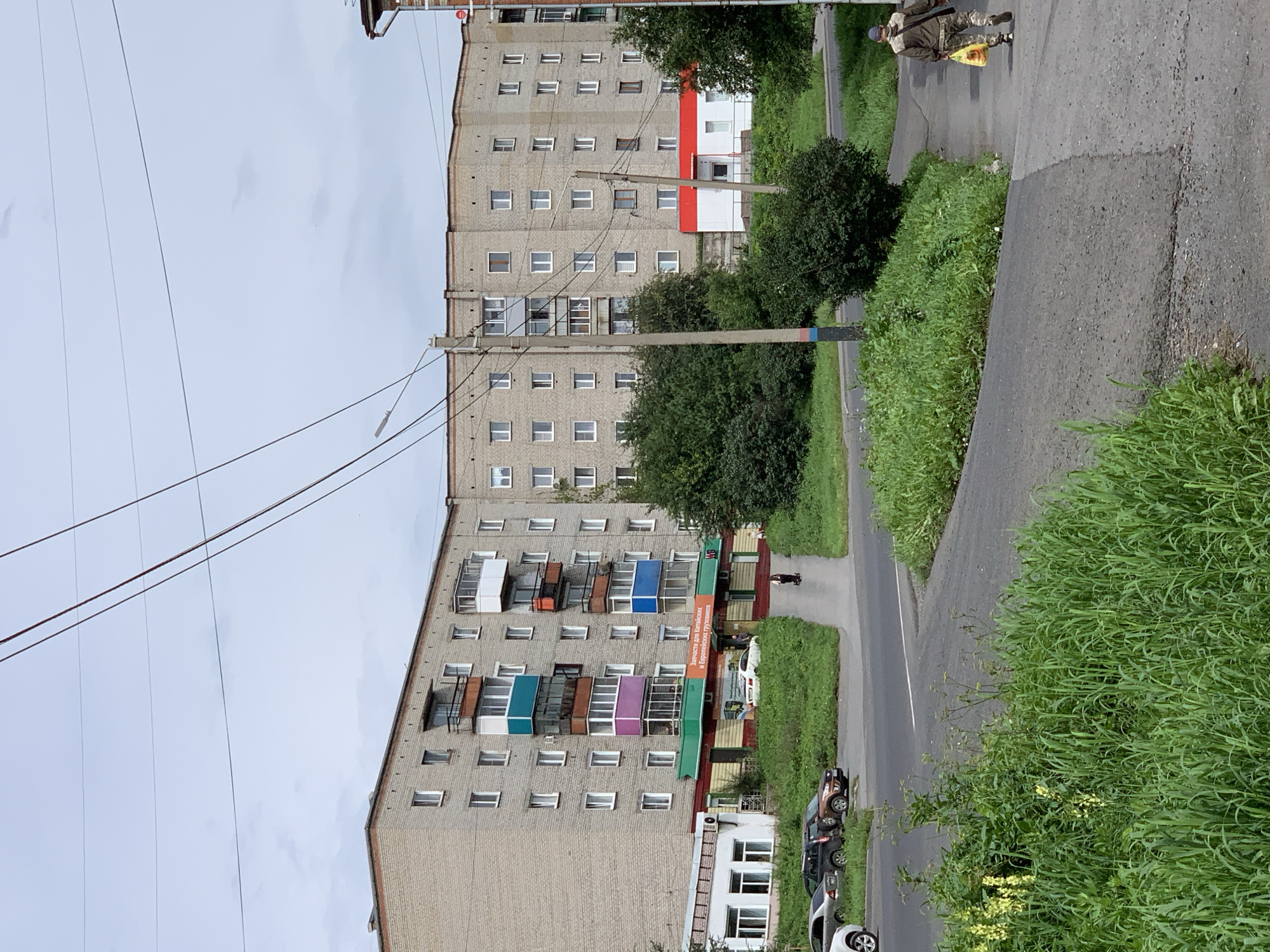 The image shows a street scene in Prokopyevsk, Russia, with a large, light-brick apartment building dominating the background. The building features multiple stories with numerous windows and balconies, some of which are painted in vibrant colors like teal, purple, and blue. A small commercial storefront is visible at the base of the building, with a sign in Cyrillic that appears to advertise auto parts. A paved road curves through the foreground, with parked cars on the left side. A man is walking away from the camera on a sidewalk on the right side of the image, carrying a yellow plastic bag. Lush green grass and trees are abundant throughout the scene, suggesting a temperate climate. The sky is overcast.