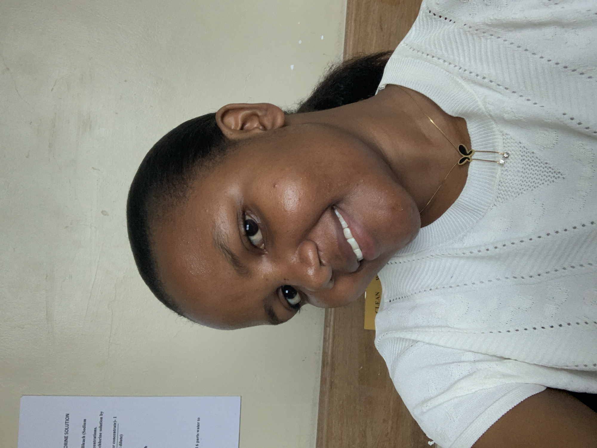A woman is positioned in front of a wall. She is wearing a white sweater and a gold necklace with a butterfly pendant. A piece of paper with the words "CORINE SOLUTION", "Bleach (Sodium", and "CLEAN" is visible in the lower left corner. The background consists of a wall and a wooden surface. There is no specific information available to determine the location of this image to be in Dar es-Salaam, Tanzania.