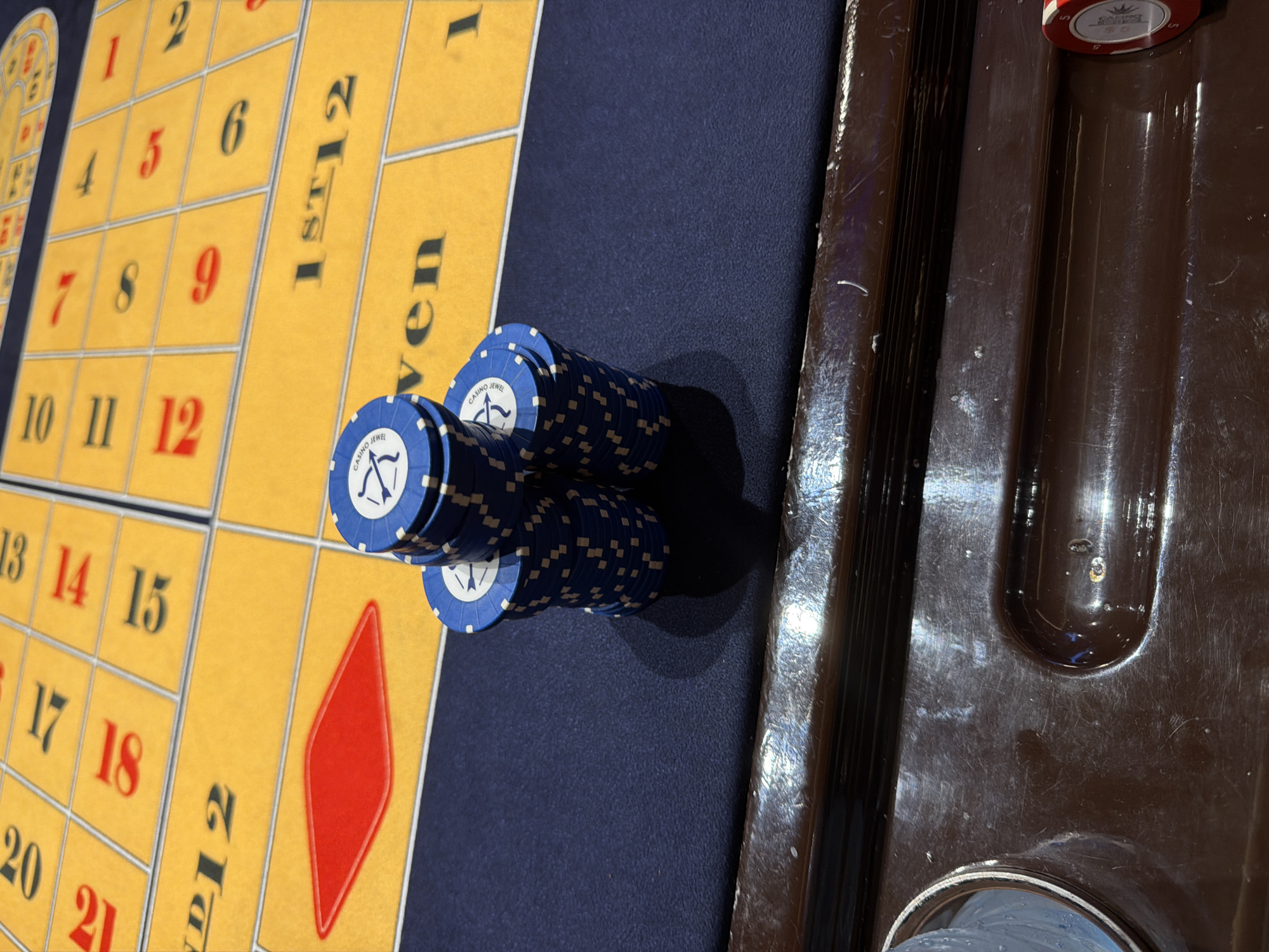 A close-up, high-angle shot captures a roulette table with stacks of blue poker chips in the foreground. The yellow felt of the table is visible, with various betting sections marked. Some numbers, like "12", "13", "14", "15", "17", "18", "20", "21", "1st 12", and "Even", are clearly legible. The chips have white and blue accents, and some bear the inscription "CASINO JR". In the lower right of the frame, a red poker chip with "5" is partially visible. The table is situated on a dark blue felt surface. The bottom of the image shows a brown, wooden surface with a reflective sheen, possibly part of a casino railing or table. There is no indication of the city of Tbilisi, Georgia, in the image.