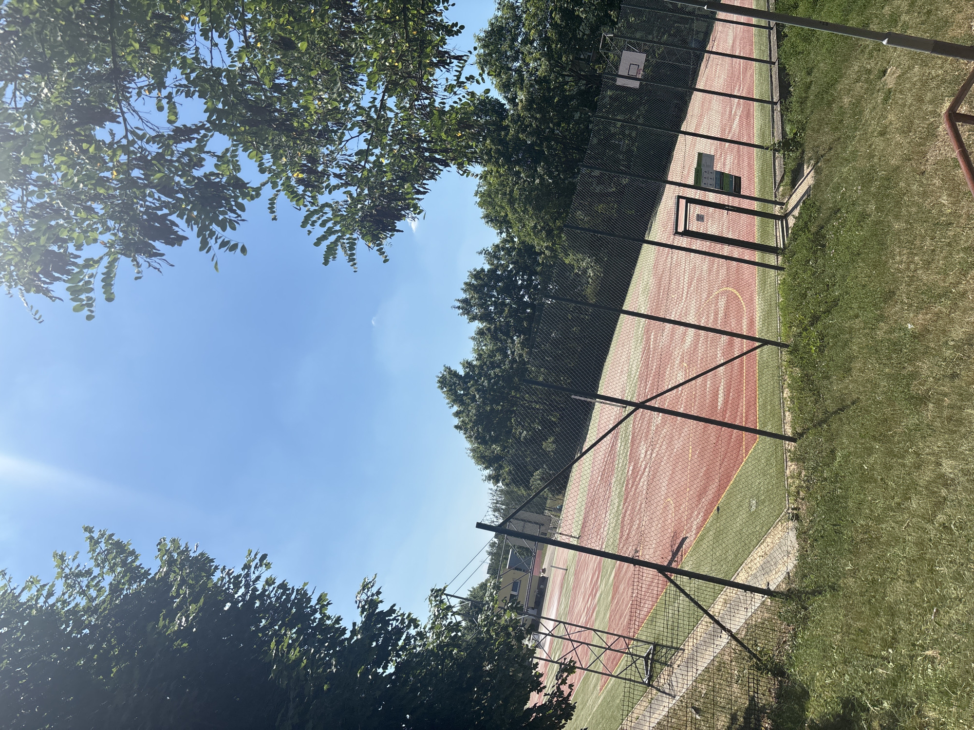This media file shows an outdoor, fenced-in basketball court on a sunny day. The court has a red and green surface with yellow lines. A basketball hoop is visible at one end of the court. Trees with green leaves surround the court, and there is also grass and a sidewalk. The sky is clear and blue. The image appears to be taken from a slightly elevated angle, looking down at the court. There is no text visible in the image. The scene suggests a recreational area, possibly a park or schoolyard. The bright sunlight indicates it is daytime.