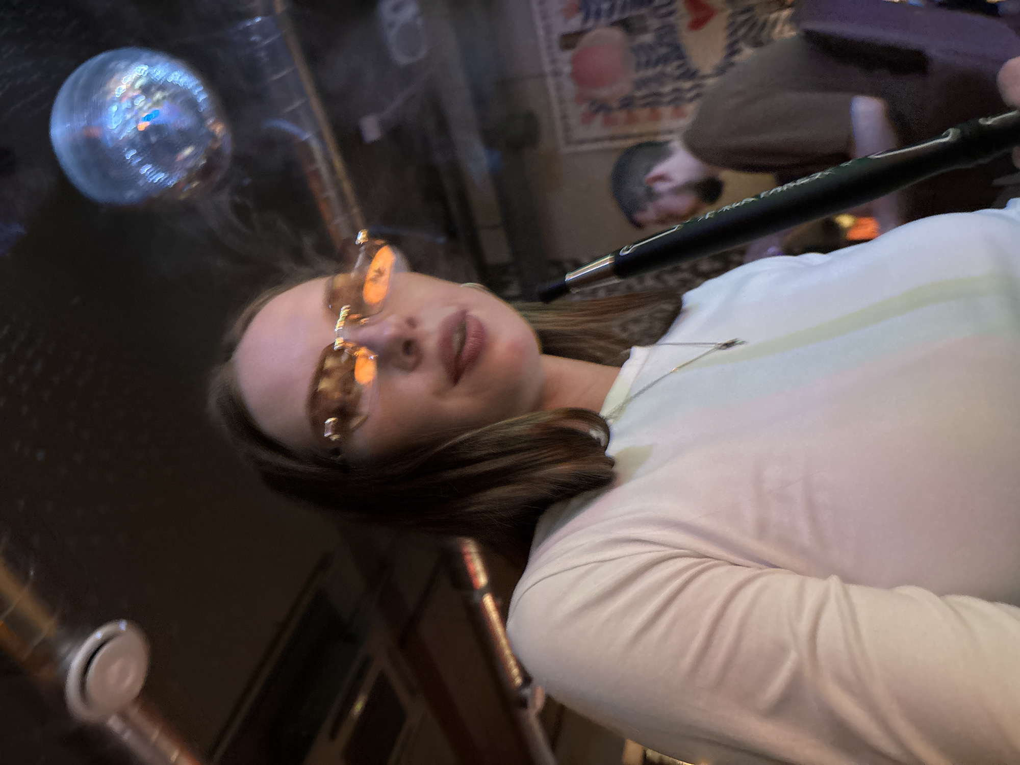 A woman with brown hair and sunglasses is holding a black hookah hose and smoking in a dimly lit room. She is wearing a white long-sleeved shirt and a silver necklace. In the background, a disco ball hangs from the ceiling, and another person is visible. The setting appears to be a hookah lounge or bar.