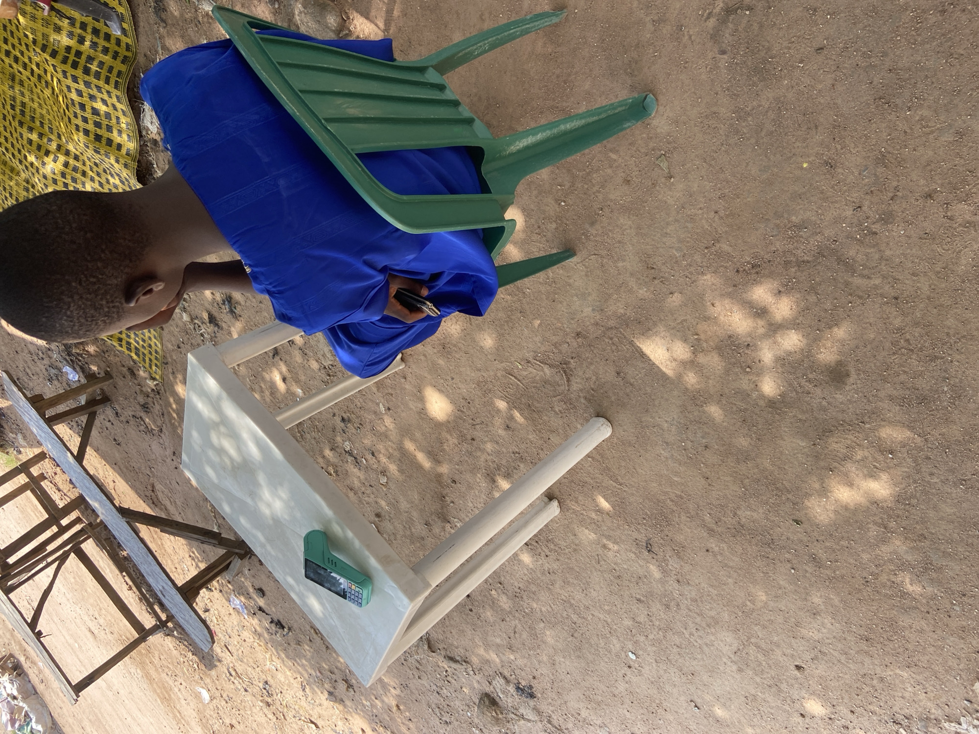 A young person, likely male based on haircut and attire, wearing a blue kaftan, sits facing away from the camera in a green plastic chair. They are seated at a white rectangular table, on which a teal-colored point-of-sale (POS) device is placed. The table has four white legs and appears to be made of plastic. The background consists of a dirt ground with scattered debris. In the distance, there is a rustic wooden table structure and a yellow and black patterned mat. The scene is outdoors, possibly in a market or street-side vending area, in Jalingo, Nigeria. The lighting suggests it is daytime, with dappled sunlight falling on the ground.
