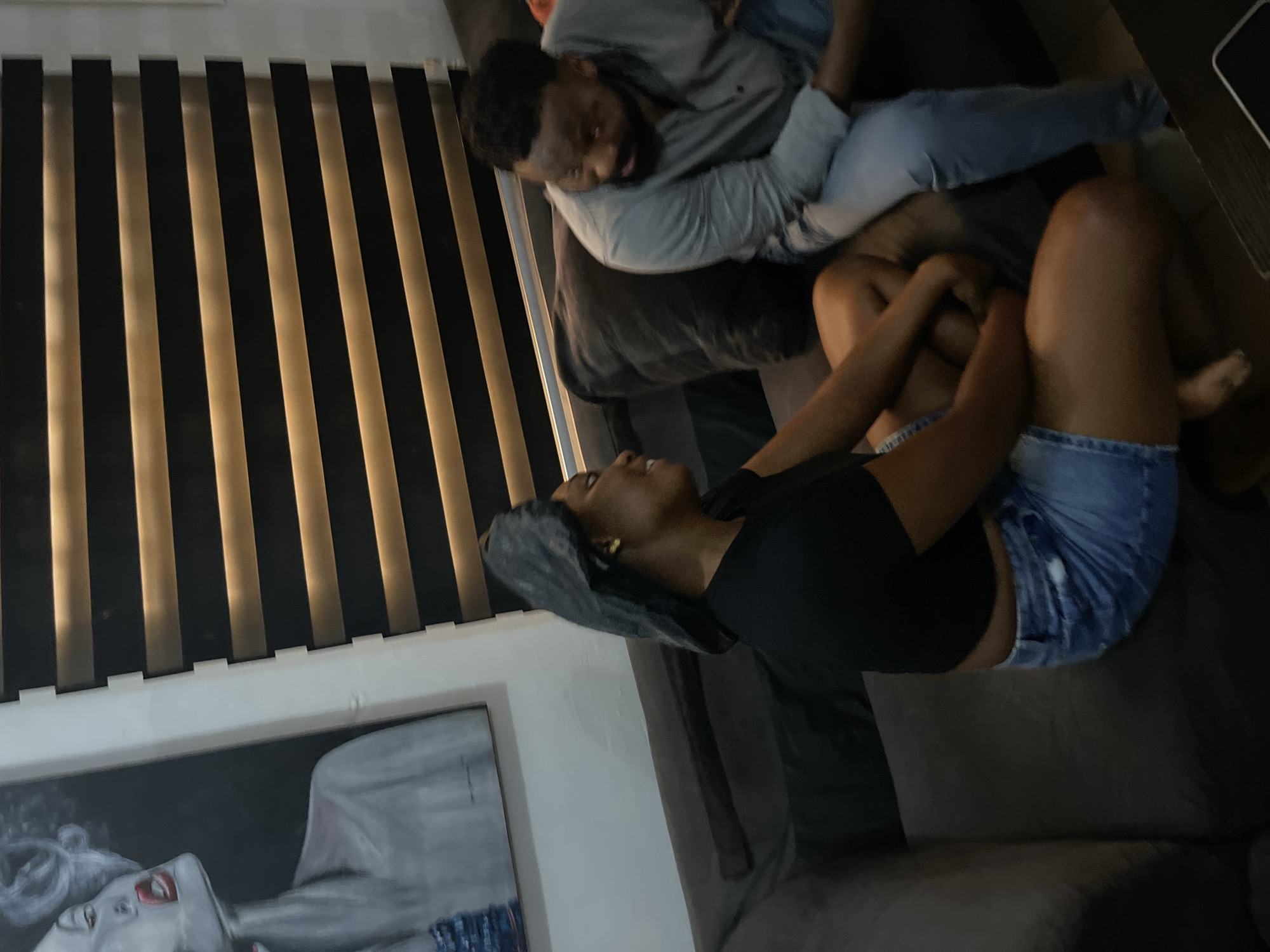 Two people are relaxing on a sofa in what appears to be a living room. The man, dressed in a light blue shirt and grey sweater, is lying down with his head resting on a pillow. The woman, wearing a black top and denim shorts, is sitting up and facing him. They seem to be engaged in a conversation, with both individuals looking at each other.
Behind them, a set of vertical blinds is visible, casting striped shadows across the room. To the left of the frame, a painting hangs on the wall, depicting a woman's face. The overall lighting suggests it might be evening or a dimly lit interior. The scene conveys a sense of casual relaxation and intimacy. The context provided indicates the location is Abuja, Nigeria. No visible text is present in the image.