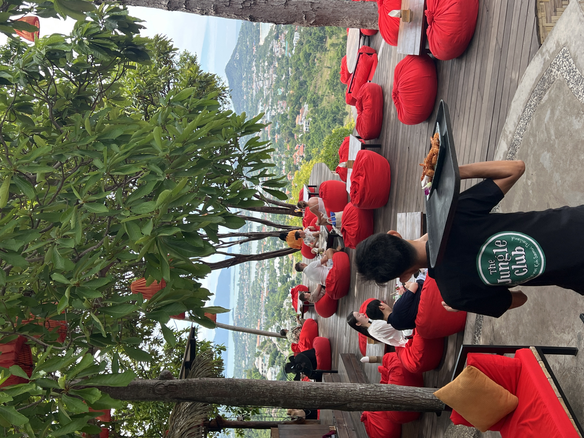 A waiter in a black t-shirt with "The Jungle Club" logo walks across a wooden deck, carrying a tray with fried food. He is in the foreground, facing away from the camera. Behind him, several patrons are seated on large, red beanbag chairs. Some are interacting with each other, while others are looking out at the view.
The setting appears to be a restaurant or bar with a scenic overlook. The deck is made of dark wood planks and is dotted with small tables. Red beanbag chairs are arranged throughout the space, some singly and others in small groups. In the background, a lush green landscape stretches out, leading to a body of water and a distant town with buildings and mountains. The scene is framed by the branches and leaves of large trees, with red lanterns hanging among the foliage. The overall atmosphere is relaxed and inviting, suggesting a casual dining experience with a beautiful view. The location is identified as Baan Chaweng Noi, Thailand.