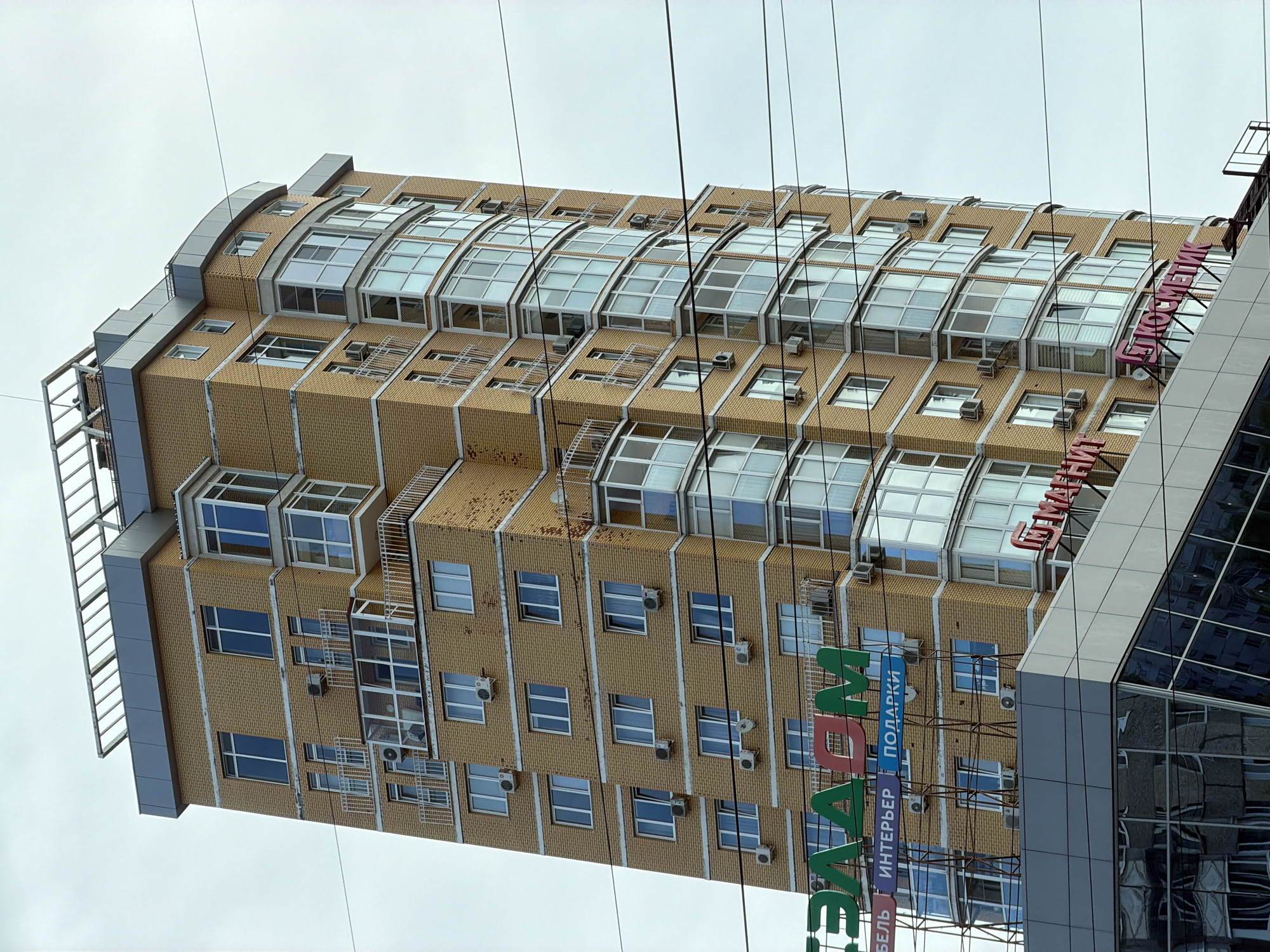 The image shows a modern, tall, yellow brick residential building in Izhevsk, Russia. The building has numerous windows and balconies, many of which are enclosed with glass. The structure curves slightly on one side, featuring a prominent bay window section. There are several signs visible on the building. One large green sign reads "ЭЛЛАДА" (Ellada), with smaller signs below it in blue and red that say "МЕБЕЛЬ" (Furniture), "ИНТЕРЬЕР" (Interior), and "ПОДАРКИ" (Gifts). Further to the right, a pink sign in cursive letters appears to say "КОСМЕТИК" (Cosmetic). The building is situated in an urban environment, with power lines crossing in front of it. The sky is overcast. Below the building, the reflective glass facade of another structure is visible, mirroring the surroundings.