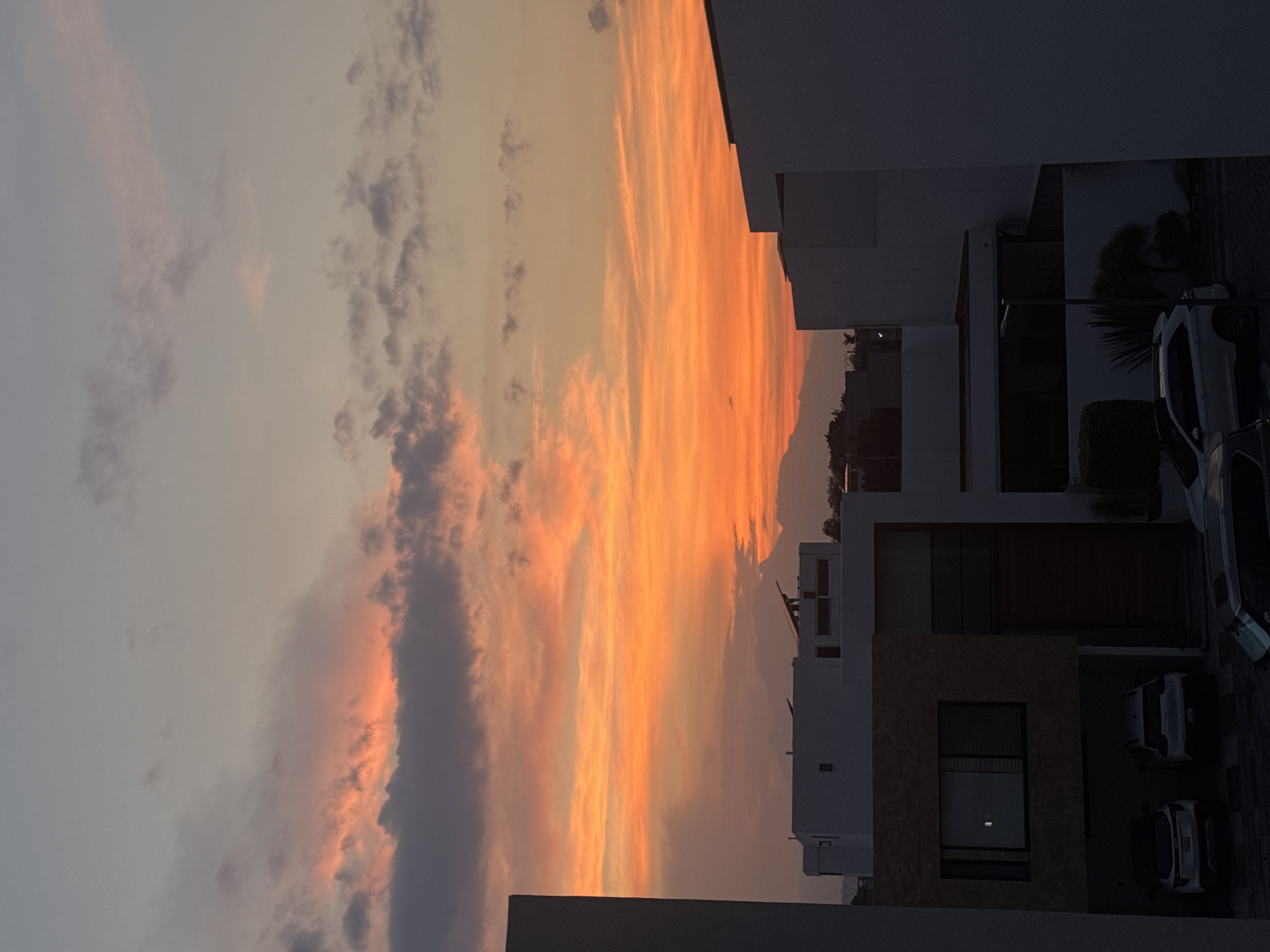 The photograph shows a sunset view from a location in Puebla, Mexico, looking east. The sky is filled with clouds ranging in color from gray to orange and pink hues, indicating a sunset. The foreground contains residential buildings, primarily white, with cars parked alongside. There are trees visible amidst the buildings. The overall lighting is dim with silhouettes cast by the buildings.