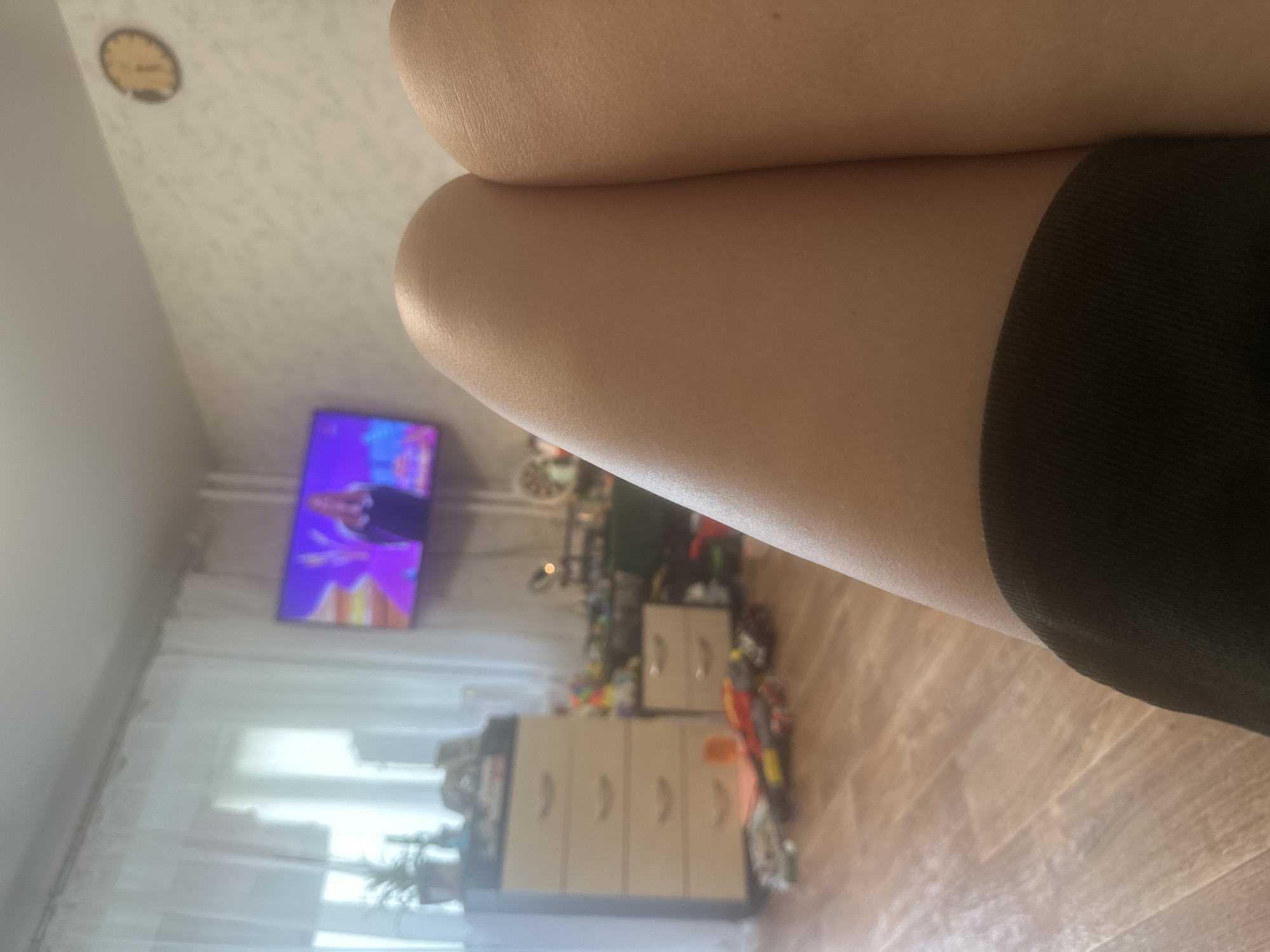 A person's legs are in the foreground of the image, with a black garment visible at the bottom. The background shows a room with a television displaying a performance, a dresser, a smaller cabinet, and toys scattered on the floor. A clock is visible on the wall. The scene appears to be an indoor living space.