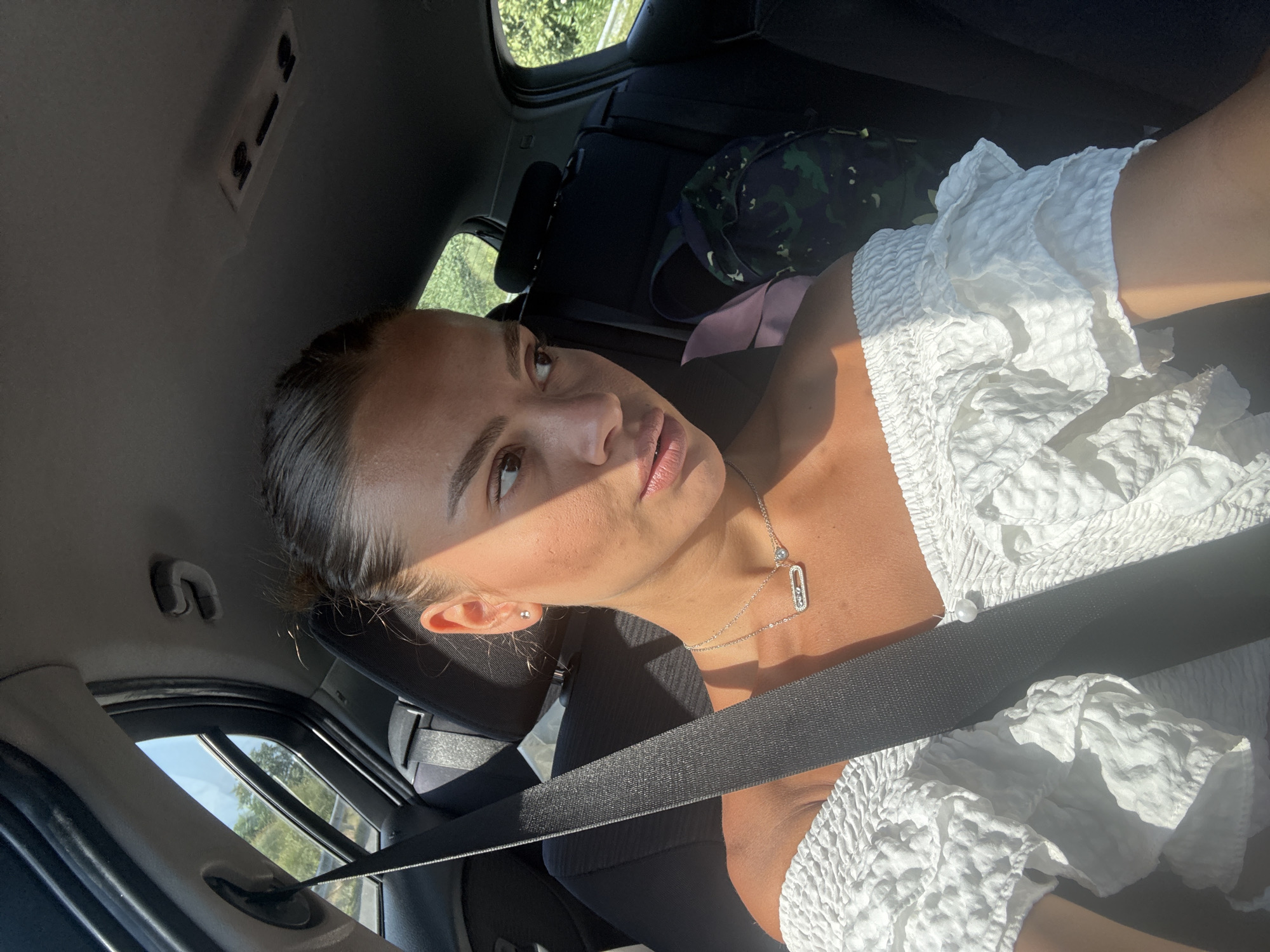 A woman with dark hair pulled back in a bun is seated in the passenger seat of a car, looking away from the camera. She is wearing a white off-the-shoulder top with ruffled sleeves and a silver necklace. Sunlight is casting a shadow across her face. A black seatbelt is visible across her chest. Outside the car window, trees and a roadside are visible, suggesting a rural or suburban setting outside of Katerini Municipality, Greece. A camouflage backpack is in the back seat.