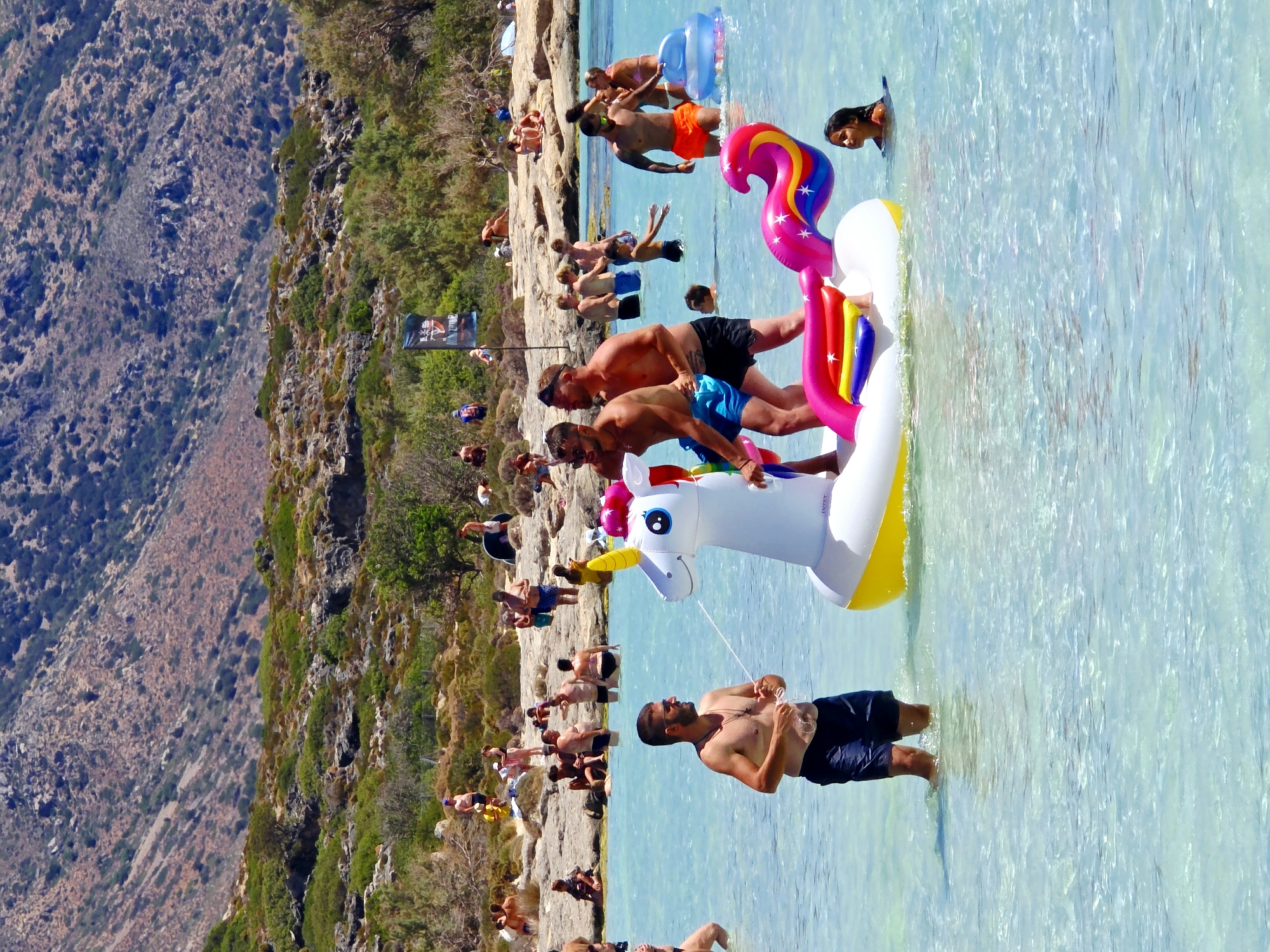 The image depicts a crowded beach scene in Elafonissi, Greece, under a clear blue sky. The foreground is dominated by the turquoise waters of the Mediterranean Sea, where several people are enjoying themselves. In the center, a large inflatable unicorn is partially submerged, with two men standing beside it. One man, wearing dark blue shorts, is holding a string connected to the unicorn's spray mechanism, while the other, in black shorts, is positioned behind him. To the right of the unicorn, a young girl with dark hair surfaces in the water. Further out, other beachgoers can be seen swimming and relaxing, some on inflatable devices. The background reveals a rocky, arid coastline with sparse vegetation and more distant figures on the shore. The overall atmosphere is one of a vibrant summer day at a popular tourist destination.
