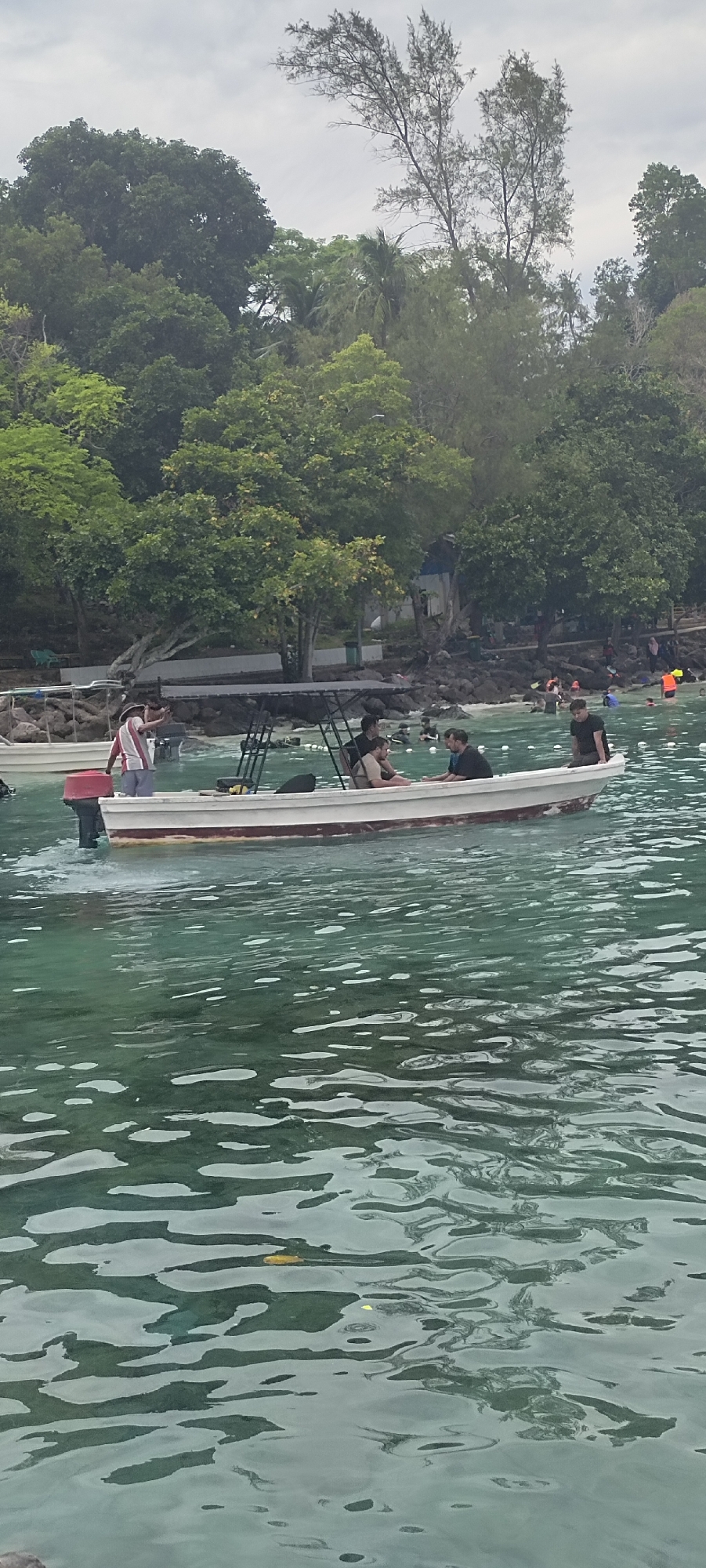 The image captures a boat with several people on board, sailing on clear, turquoise water near the shore of Sabang, Indonesia. The boat, a white vessel with a red stripe along its waterline, is equipped with a canopy. Four individuals are visible inside the boat. One person, wearing a striped shirt and shorts, stands at the stern, appearing to be directing or interacting with others. Three other individuals are seated in the boat, one wearing a light-colored shirt and dark shorts, another in a dark t-shirt, and a third in a black t-shirt. In the background, a lush green, tree-covered hillside slopes down to a rocky coastline. A few other boats are visible further out in the water, and several people are swimming or wading near the shore, some wearing life jackets. The sky is overcast, suggesting a cloudy day.