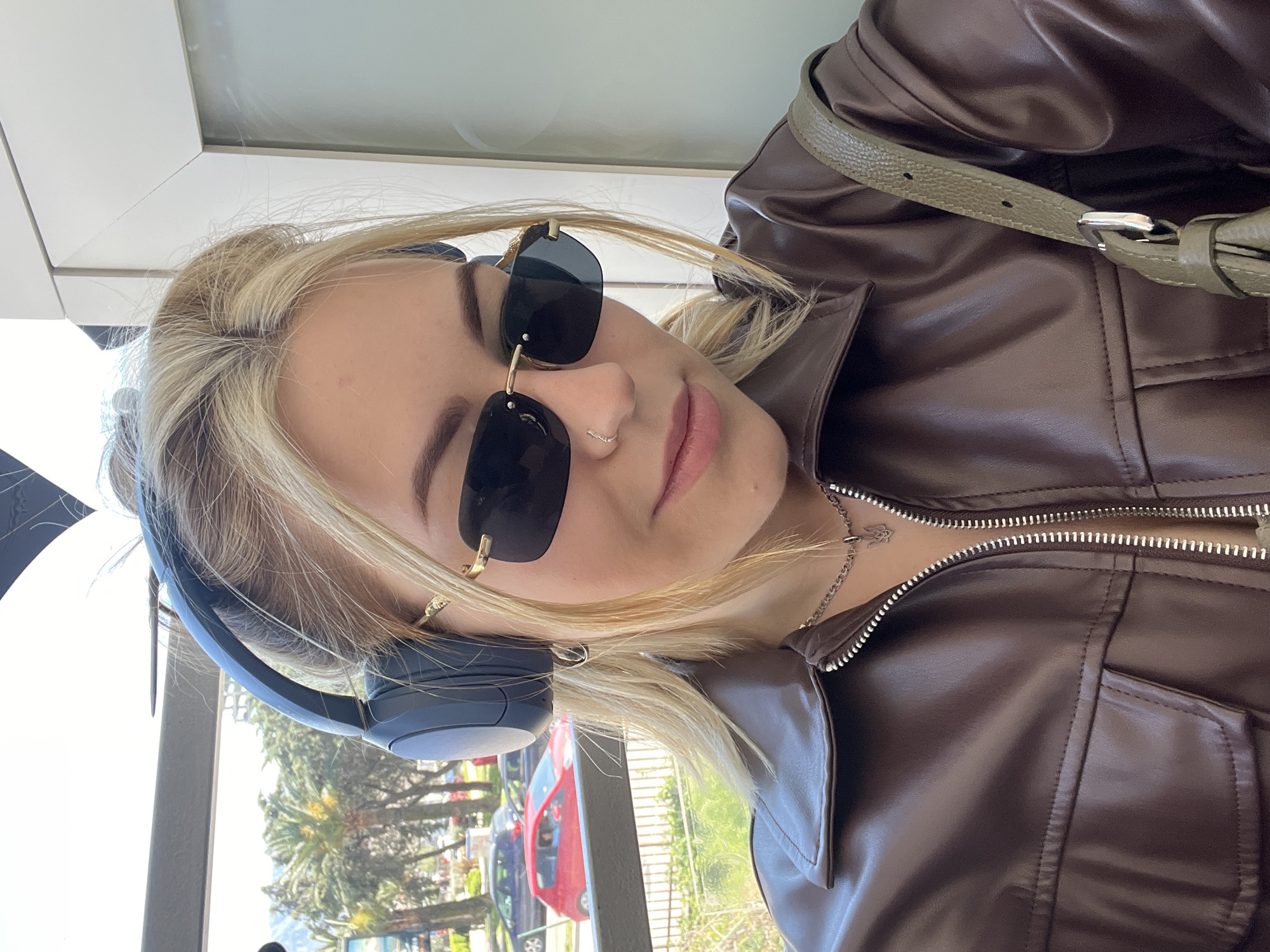A young, blonde woman wearing dark sunglasses and headphones is indoors, likely near a window. She has a nose ring and is wearing a brown, zip-up leather jacket with a green purse strap visible. Outside the window are palm trees and vehicles, including a red car, suggesting an urban setting. This image was taken in Budva, Montenegro.