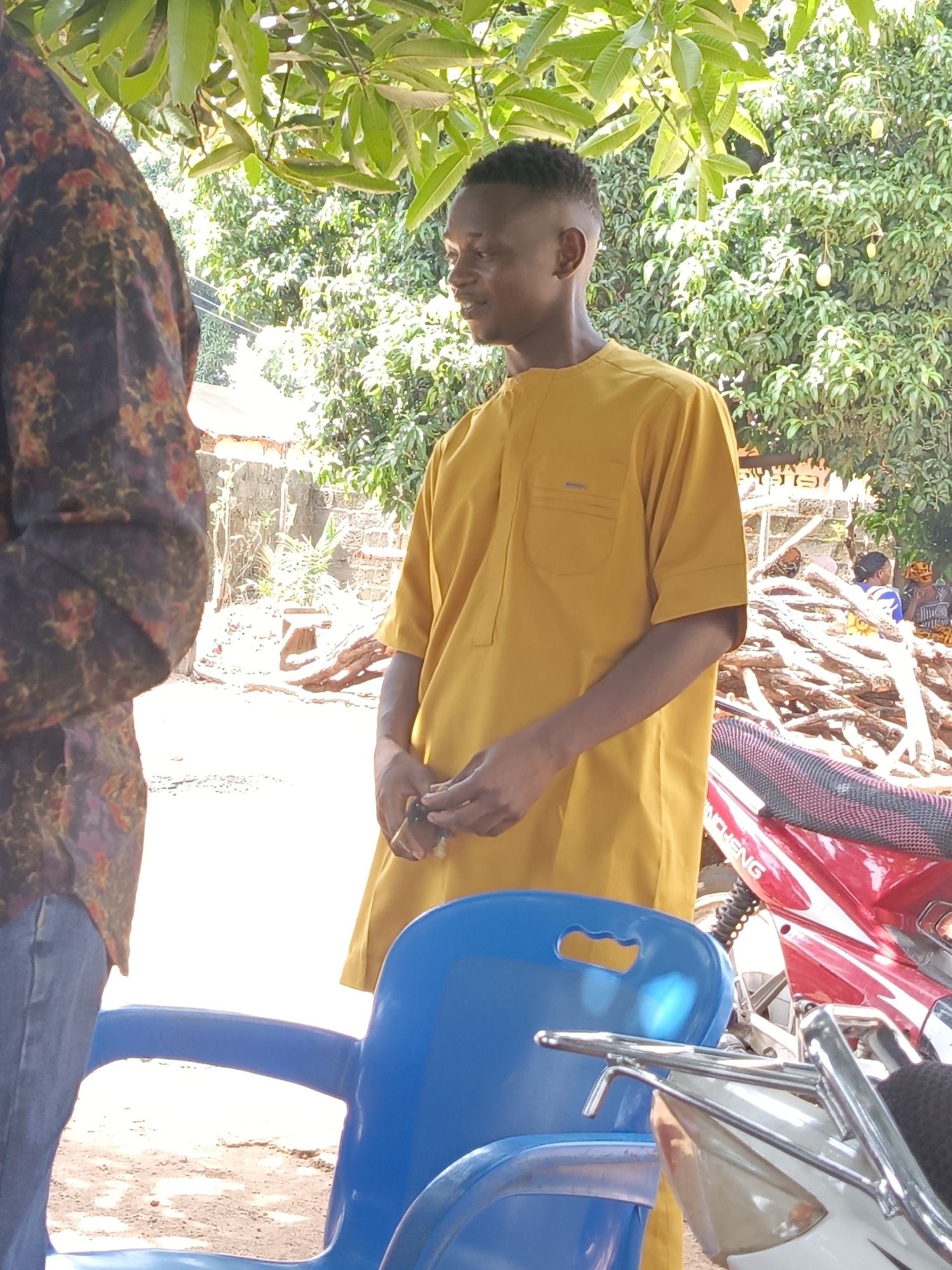 In an outdoor scene in Akwanga, Nigeria, a man in a mustard-colored, short-sleeved garment stands next to a blue plastic chair and a parked red and white motorcycle. Another person, partially visible, wears a patterned shirt and jeans. Behind the main subject, a pile of firewood is stacked near individuals in traditional clothing. Overhanging foliage provides partial shade.