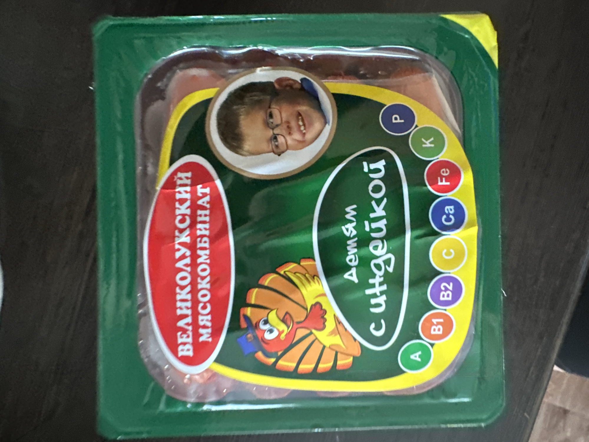 The image shows a close-up, overhead view of a package of food products, likely sausages or cured meats, presented in a green plastic tray with a clear plastic film cover. The packaging is predominantly green and yellow, with various labels and illustrations.
A prominent label on the left side is oval-shaped and red, with white Cyrillic text that reads "ВЕЛИКОЛУКСКИЙ МЯСОКОМБИНАТ" (Velikoluksky Meat Combine).
In the center of the packaging, a yellow curved band contains white Cyrillic text that appears to be "детям сгущёной" (for children condensed). Below this text is a cartoon illustration of a bird wearing a blue hat, with red and yellow features, set against a background of radiating orange lines.
To the right of the central text, a series of colorful circles with letters inside represent vitamins or nutrients. These circles are arranged vertically and include: 'P' (blue), 'K' (green), 'Fe' (red), 'Ca' (blue), 'C' (yellow), 'B2' (purple), 'B1' (orange), and 'A' (green).
Above the central text, a circular inset displays a photograph of a young boy wearing glasses and smiling. The boy appears to be of European descent.
The packaging is placed on a dark, textured surface, possibly a wooden table, which suggests an indoor setting. The lighting appears to be natural or from an overhead source, casting subtle shadows. The overall impression is of a product packaging designed to appeal to consumers, with elements highlighting nutritional content and a child-friendly image. The location context provided is Novoaltaysk, Russia, which aligns with the Cyrillic text on the packaging.