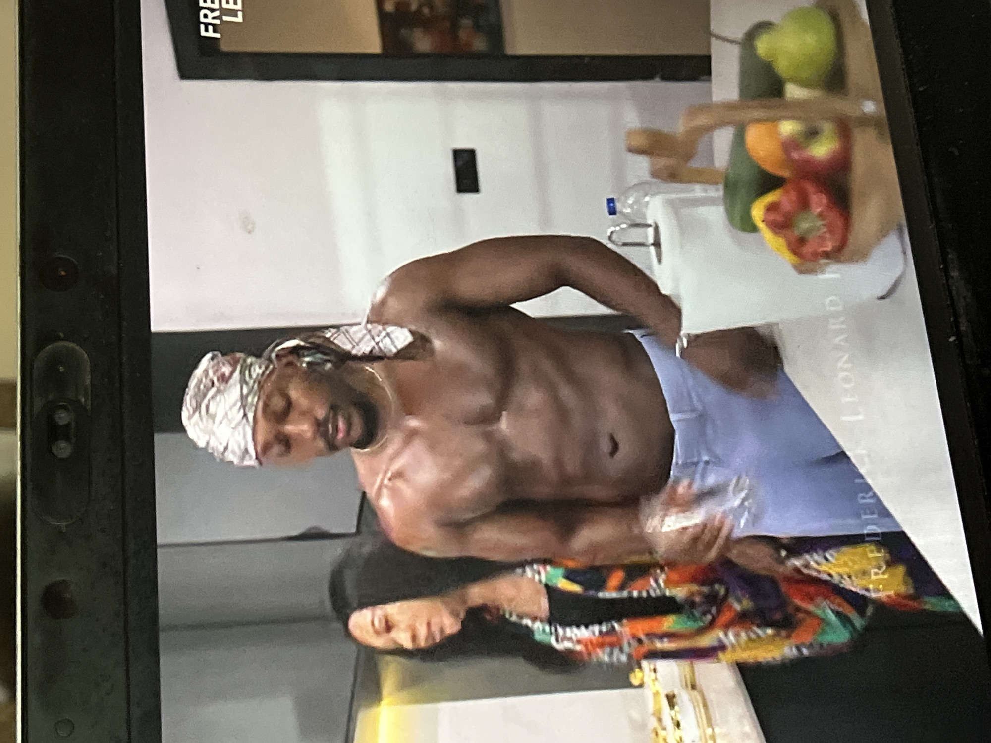 The image displays a photo taken of a screen, showing two individuals in an indoor setting, likely a kitchen or bar area. The scene is oriented vertically in the provided image but appears to be horizontally composed, showing a man and a woman.
The prominent subject is a dark-skinned, shirtless man with a muscular physique, wearing light-colored shorts or trousers and a white and grey patterned head wrap (possibly a durag or scarf). He also wears a thin gold-colored necklace. He is looking downwards or slightly to his right, with his mouth slightly ajar, suggesting he might be speaking or focused on an activity.
Below him, partly visible as a reflection, is a dark-skinned woman with long dark hair, wearing a colorful, patterned long-sleeved top or dress. Her face is seen in reflection, looking subtly upwards.
The setting includes a light-colored countertop. On the countertop, behind the man, there is a brown wooden basket or stand containing various fruits and vegetables, including a green lime or avocado, an orange, a red bell pepper, and a yellow bell pepper. A clear plastic water bottle stands near the produce.
In the background, above the counter, a dark-framed object is visible on a wall. Partially visible text on this frame reads "FRE" and "LE", likely part of a name. Additional text is visible on the screen itself, near the lower right portion of the image, which reads "FREDERICK LEONARD" as a watermark or onscreen text.
The overall impression is an intimate or casual indoor scene, possibly a still from a video or show, given the screen capture nature and visible text. The time of day is not explicitly discernible, but the lighting suggests an interior setting, possibly illuminated by artificial lights. No specific location cues point to Abuja, Nigeria, beyond the subjects' apparent ethnicity.